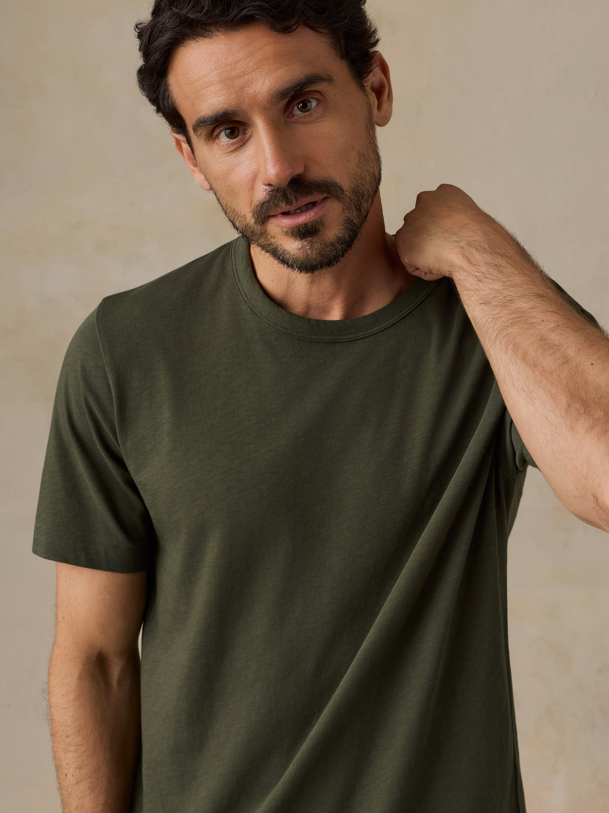 A man wearing a solid olive green short-sleeve crewneck t-shirt, posing with one hand on his neck against a neutral beige textured background., image 5 of 6