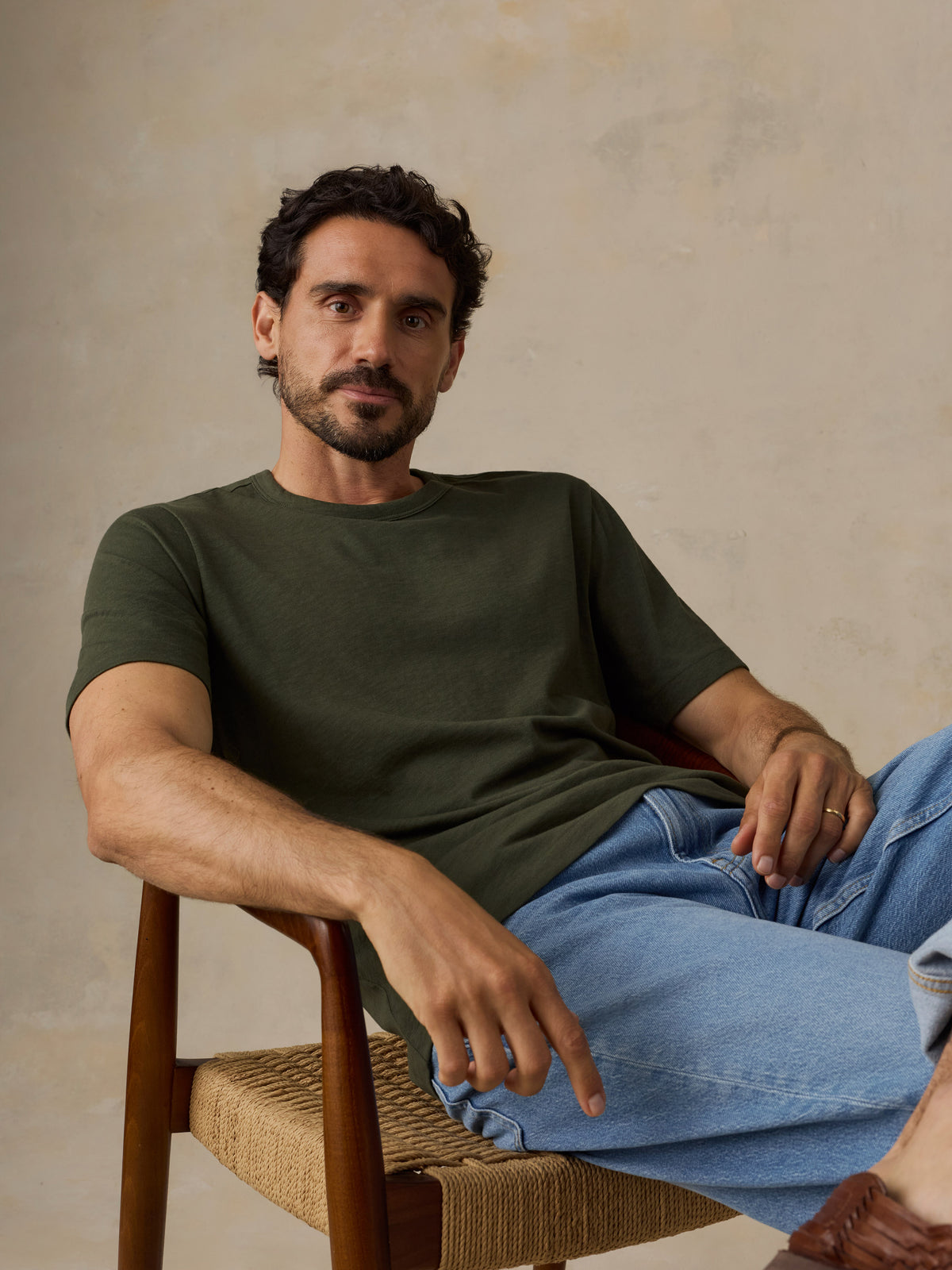 A man wearing an olive green crew-neck t-shirt and light blue jeans, sitting in a wooden chair with a woven seat against a textured beige wall., image 2 of 6