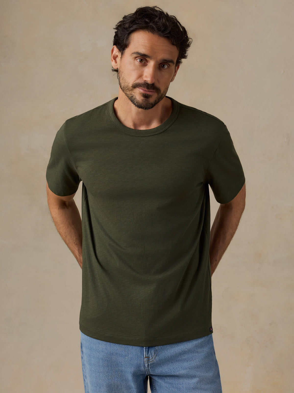 A man wearing a dark olive green short-sleeve crewneck t-shirt and light blue jeans, standing against a textured beige wall., image 1 of 6