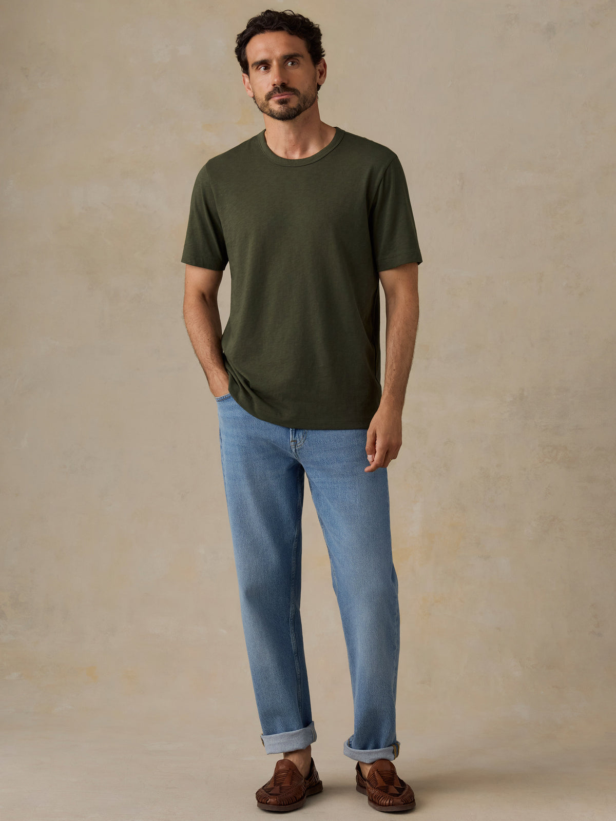 A man wearing a dark olive green crew-neck t-shirt, light blue jeans, and brown leather loafers, standing against a textured beige studio backdrop., image 4 of 6