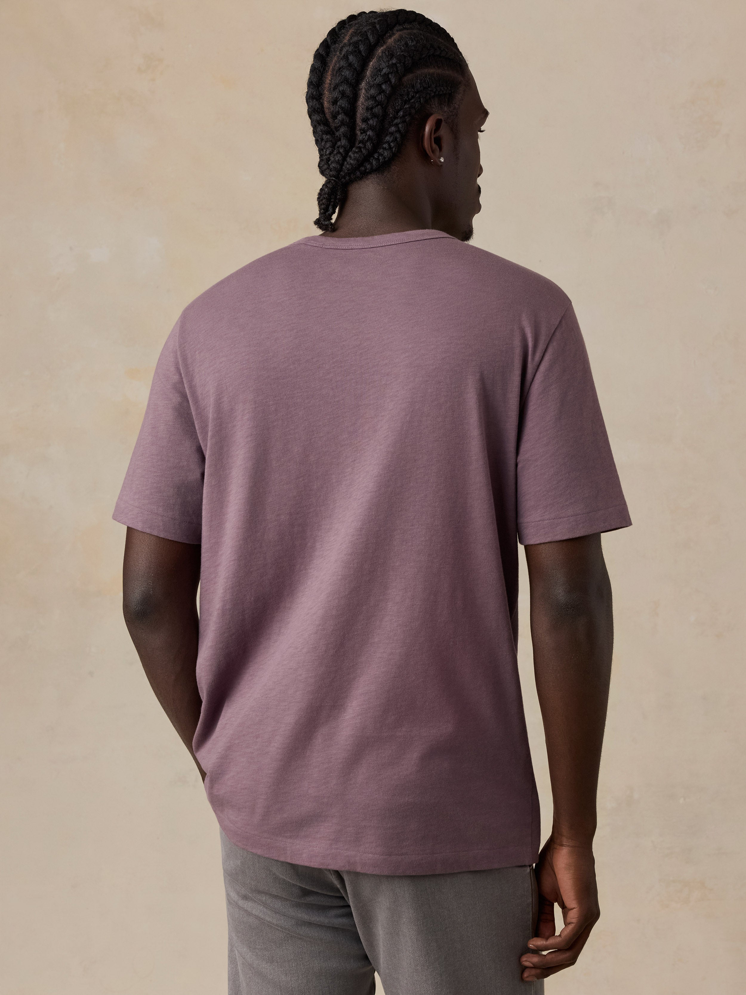 A man viewed from behind wearing an oversized, short-sleeve crewneck t-shirt in a dusty mauve-purple color, paired with gray pants, against a textured beige background.