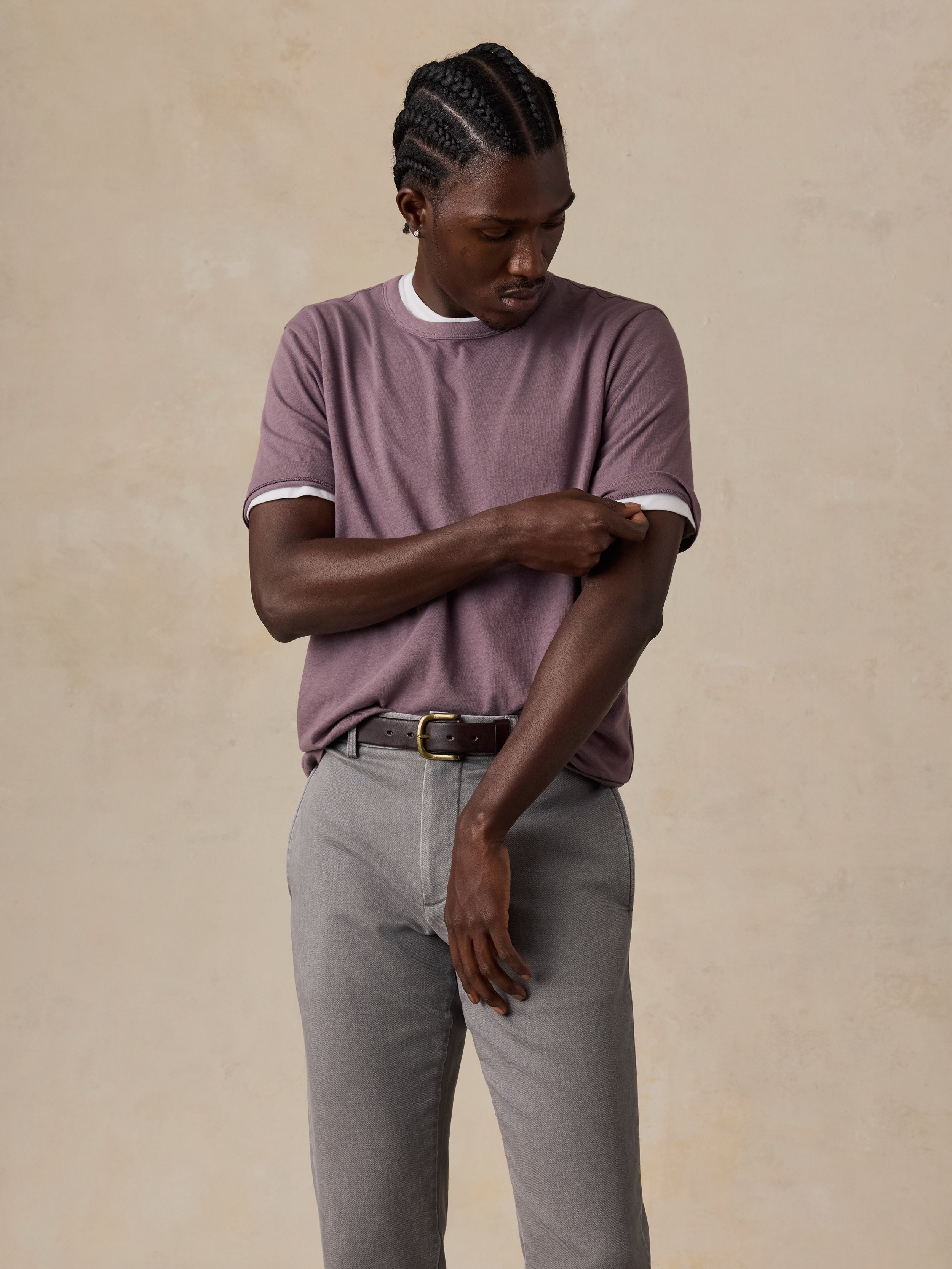 A man wearing a mauve-purple t-shirt paired with gray trousers and a dark belt, standing against a textured beige background.
