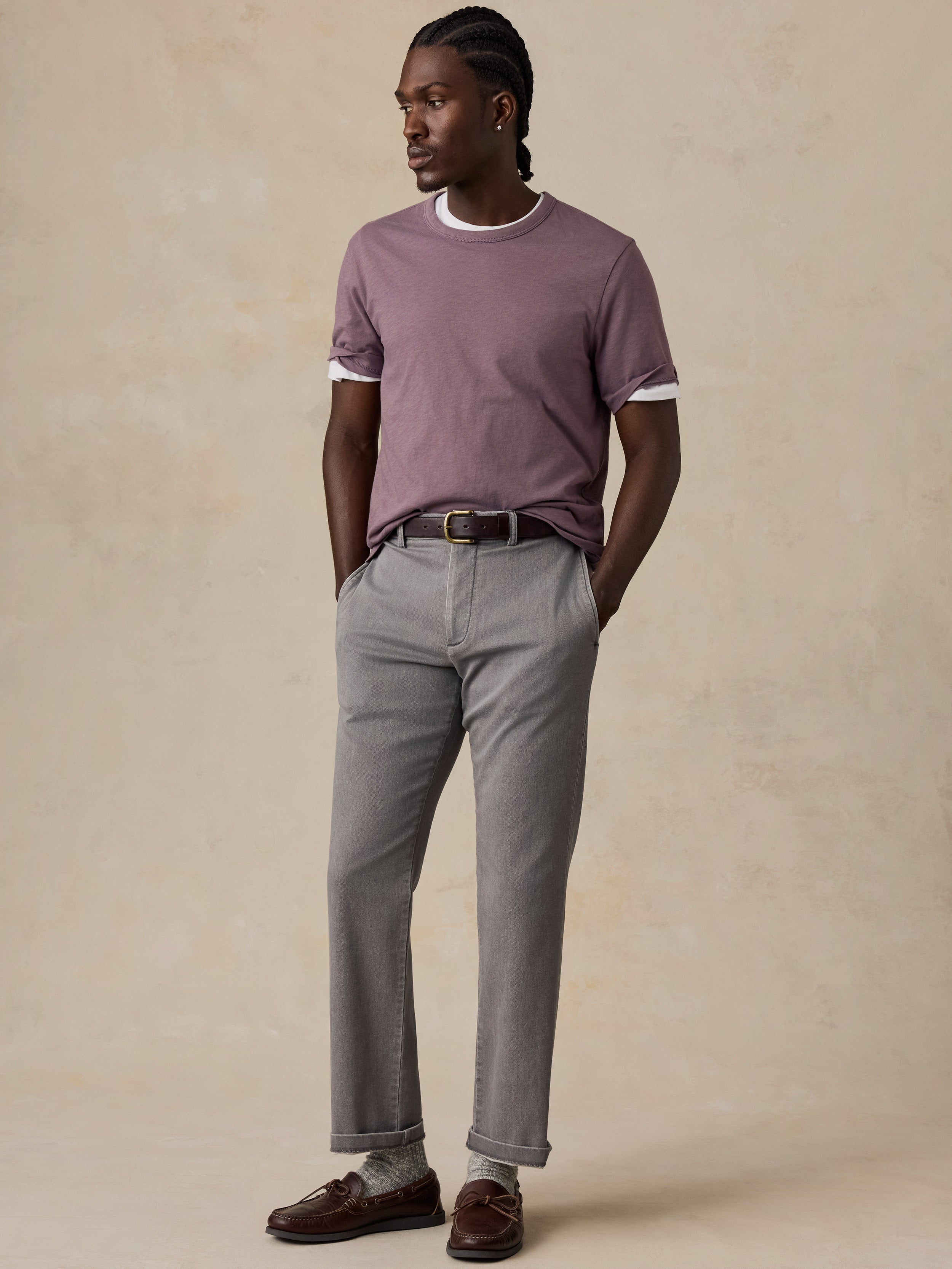 A man wearing a mauve t-shirt, gray dress pants, a dark brown leather belt, and brown leather loafers, standing against a textured beige studio backdrop.