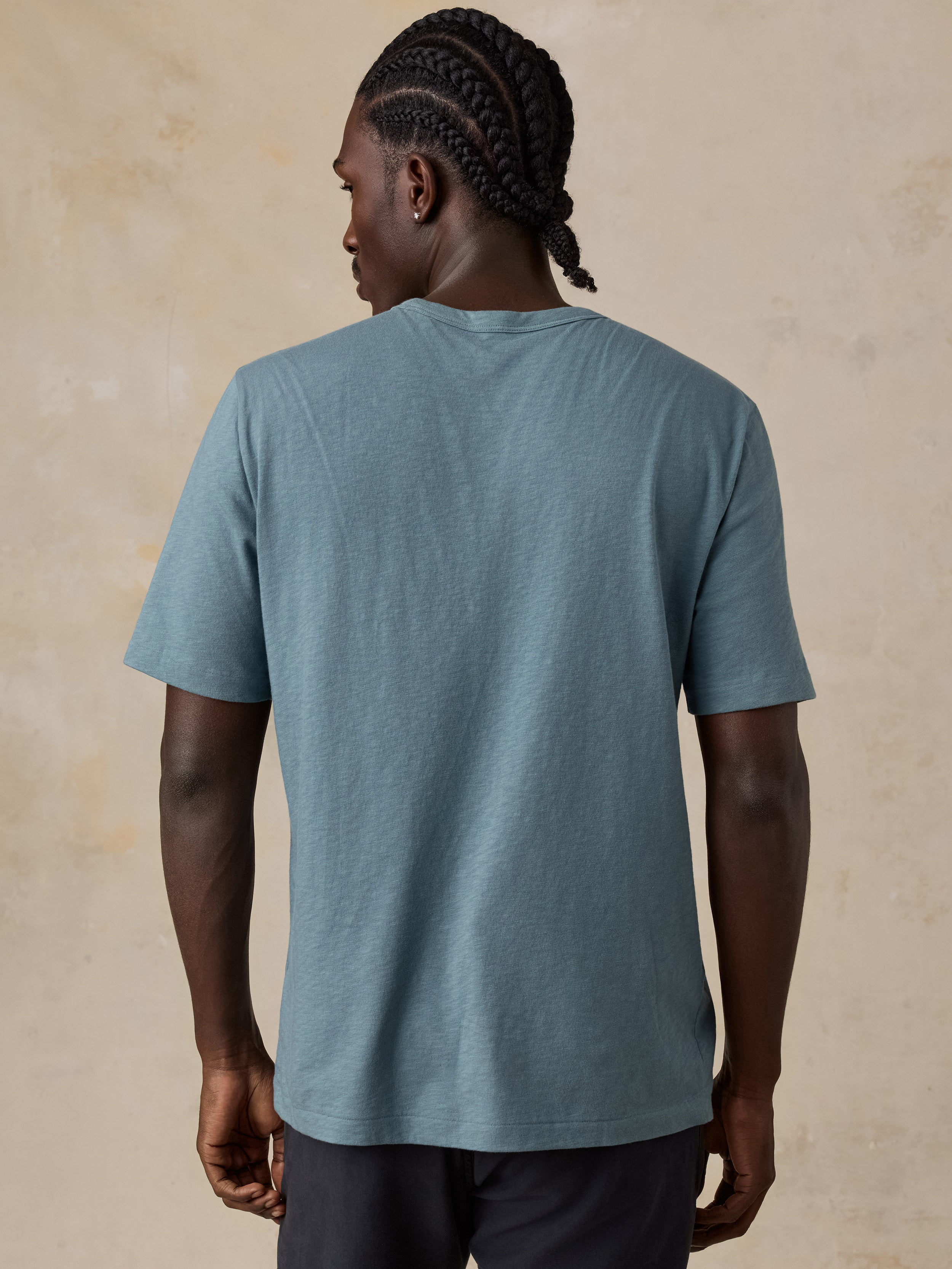 A man viewed from behind wearing a heathered slate blue crewneck t-shirt and dark pants, standing against a textured beige wall.