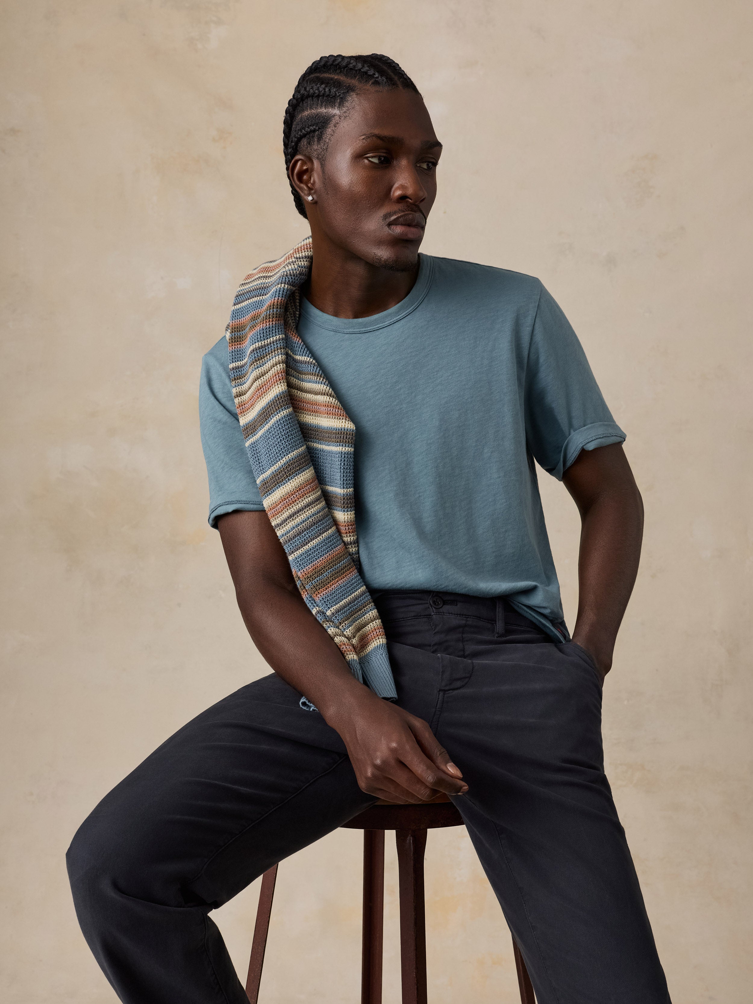 A man seated on a stool wearing a muted blue crew-neck t-shirt, dark charcoal trousers, and a striped scarf draped over his shoulder, posed against a textured beige studio backdrop.