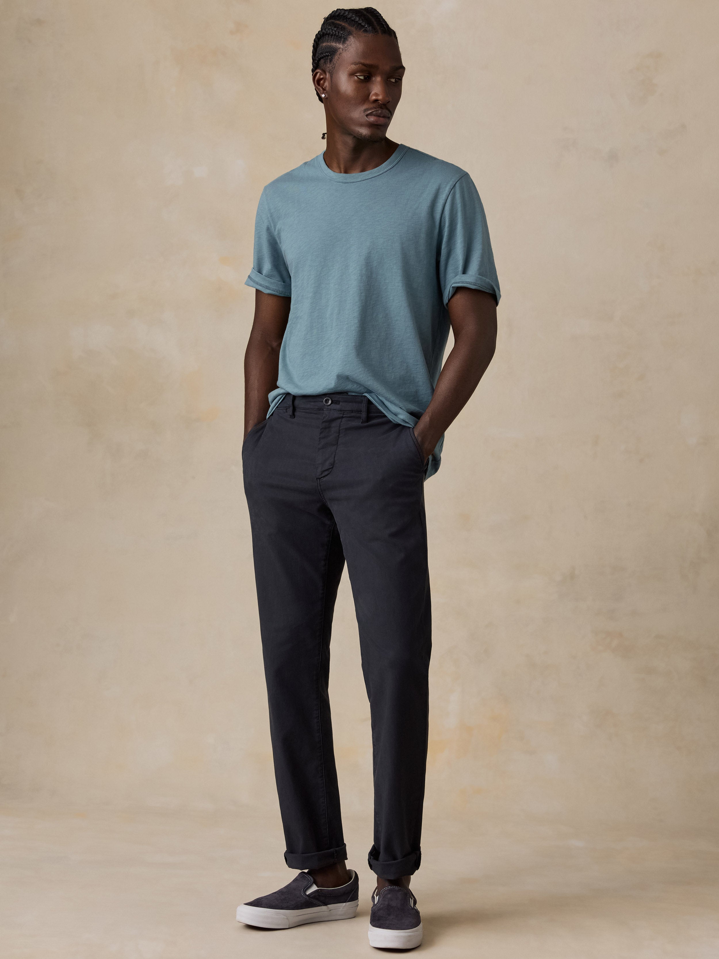 A man modeling a light blue crewneck t-shirt, dark charcoal gray slim-fit pants, and dark slip-on sneakers against a textured beige studio backdrop.
