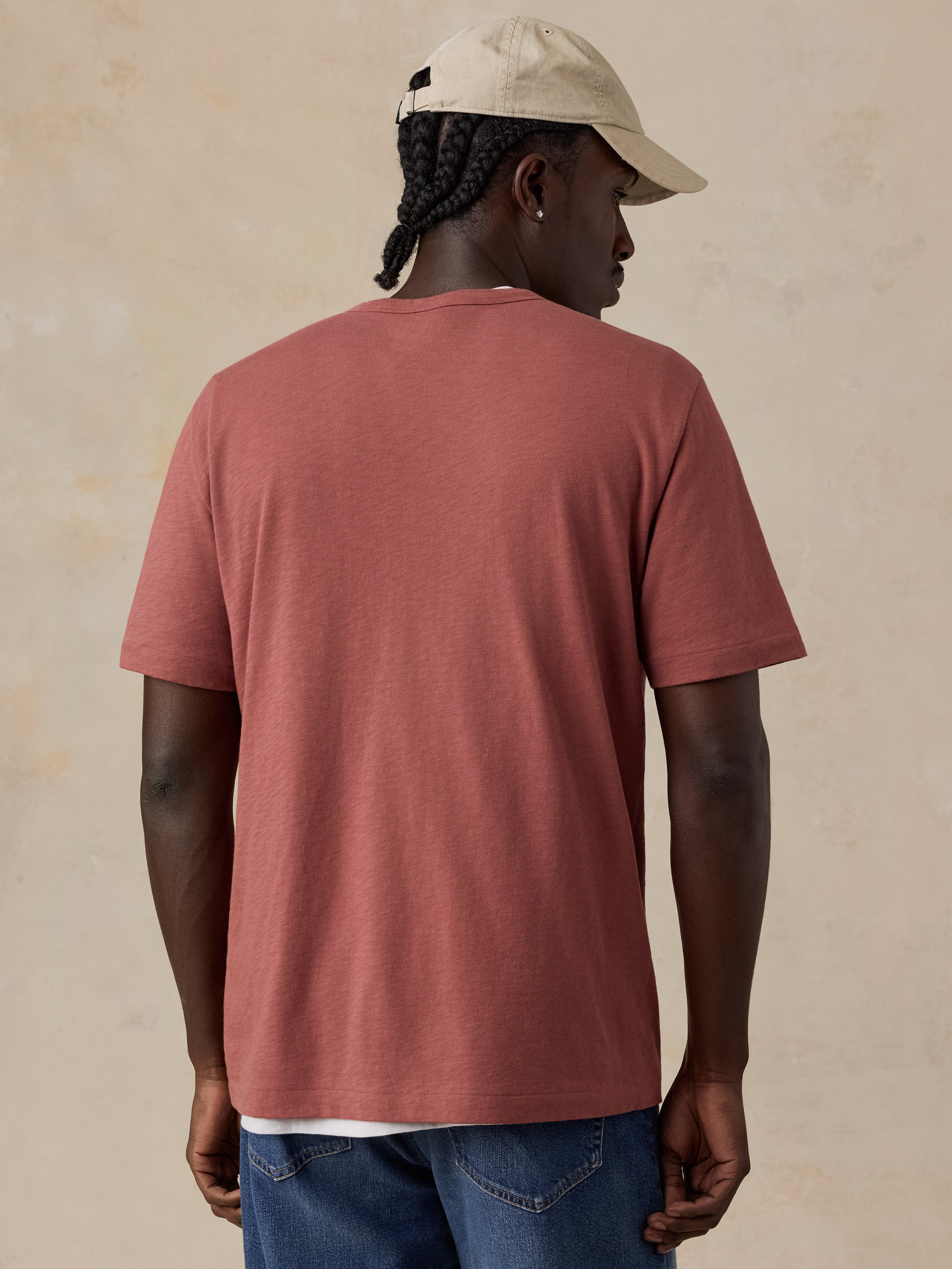 A man viewed from behind wearing a muted dusty rose short-sleeve t-shirt, blue jeans, and a beige baseball cap against a textured beige background.