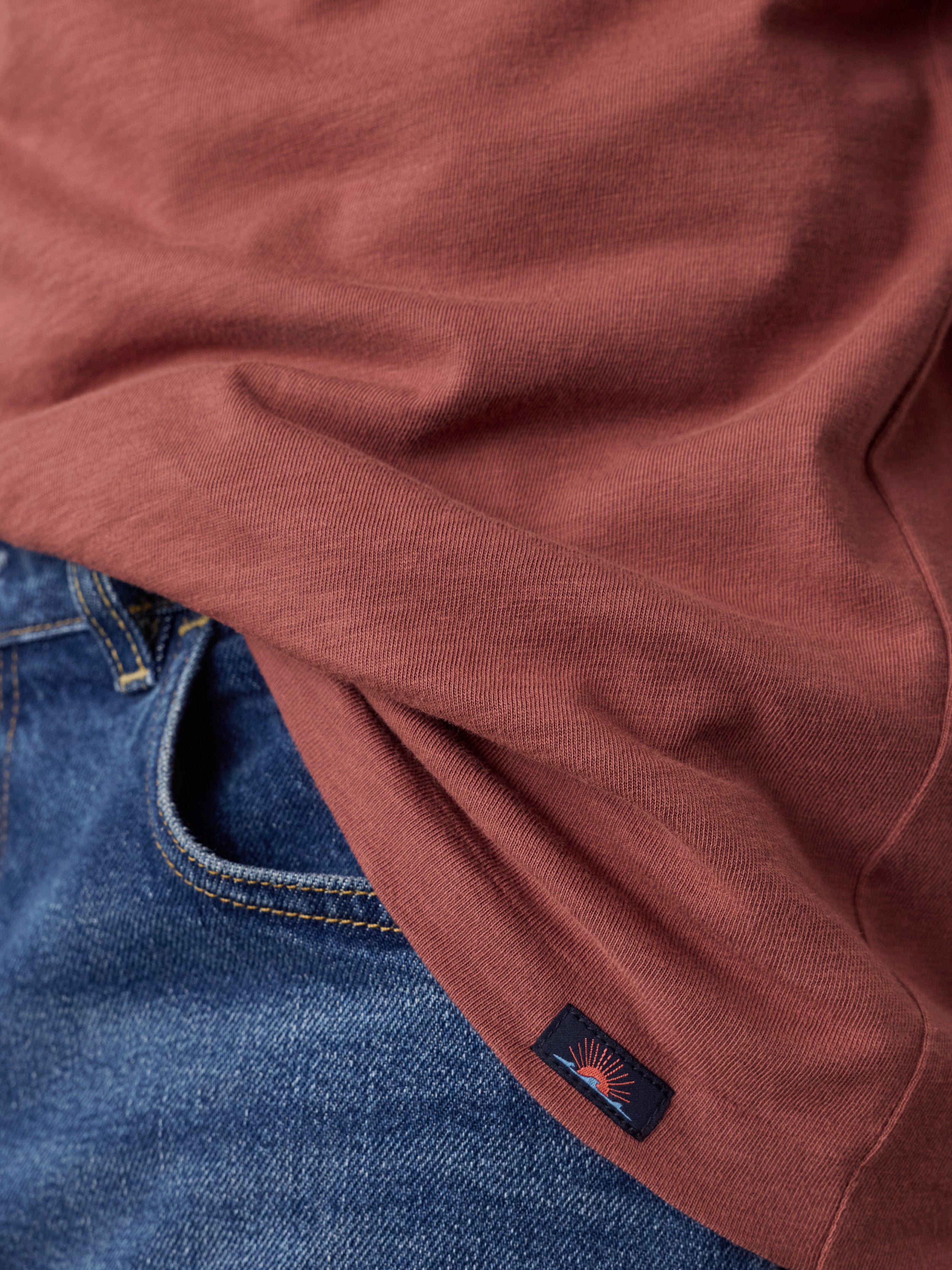 Close-up of a rust-colored t-shirt draped over blue denim jeans, showing a small Faherty brand patch near the hem