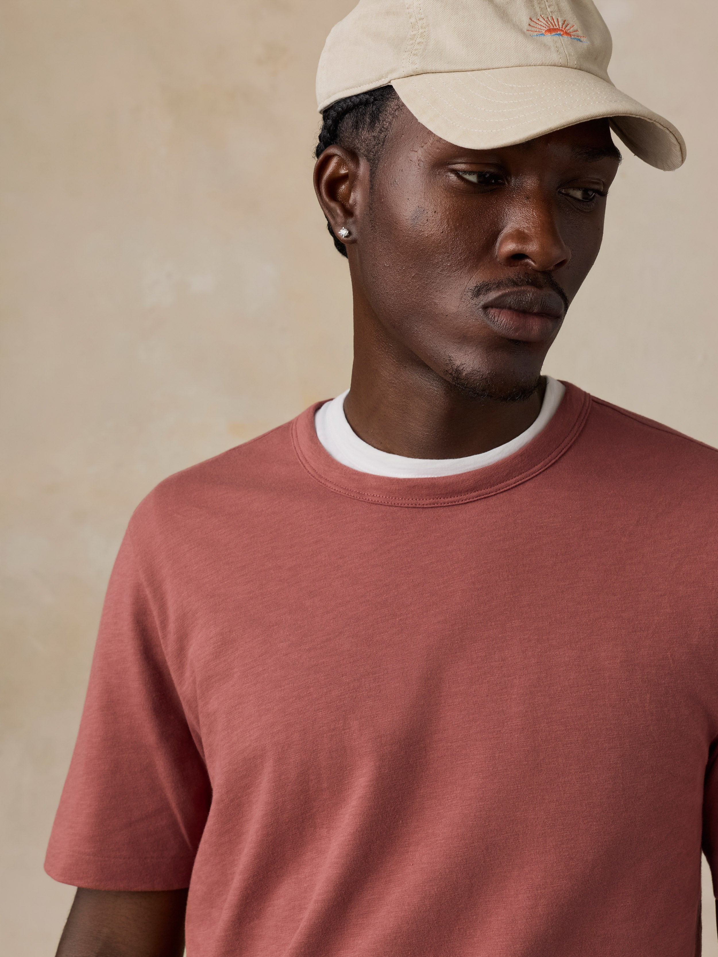 A man wearing a dusty rose crewneck t-shirt with a white ribbed collar and a beige baseball cap, photographed against a textured beige background.