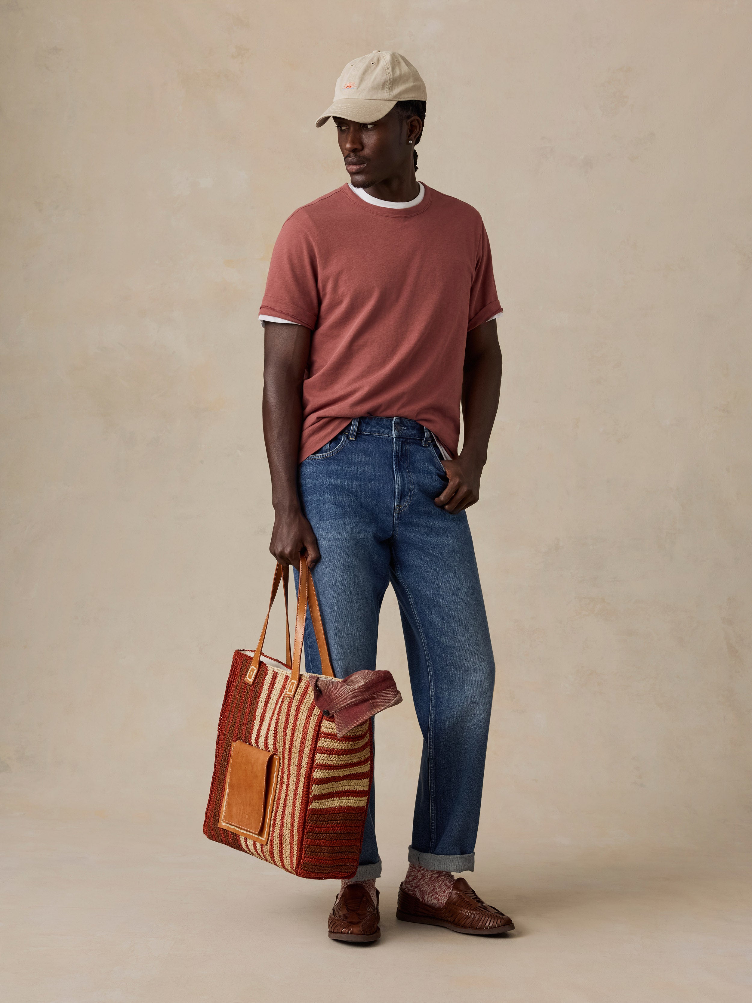 A man wearing a beige baseball cap, a dusty rose ringer t-shirt, blue jeans, and brown leather loafers stands against a textured beige studio backdrop, holding a striped woven tote bag.