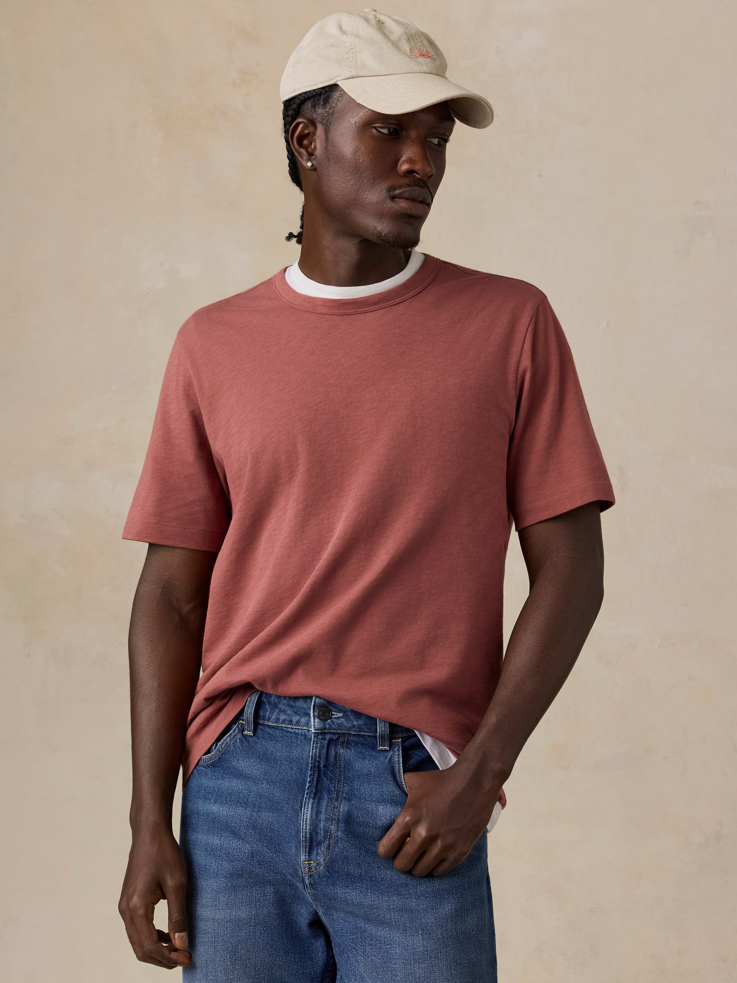 A man wearing a dusty rose short-sleeve crewneck t-shirt, blue jeans, and a beige baseball cap, standing against a textured beige background.