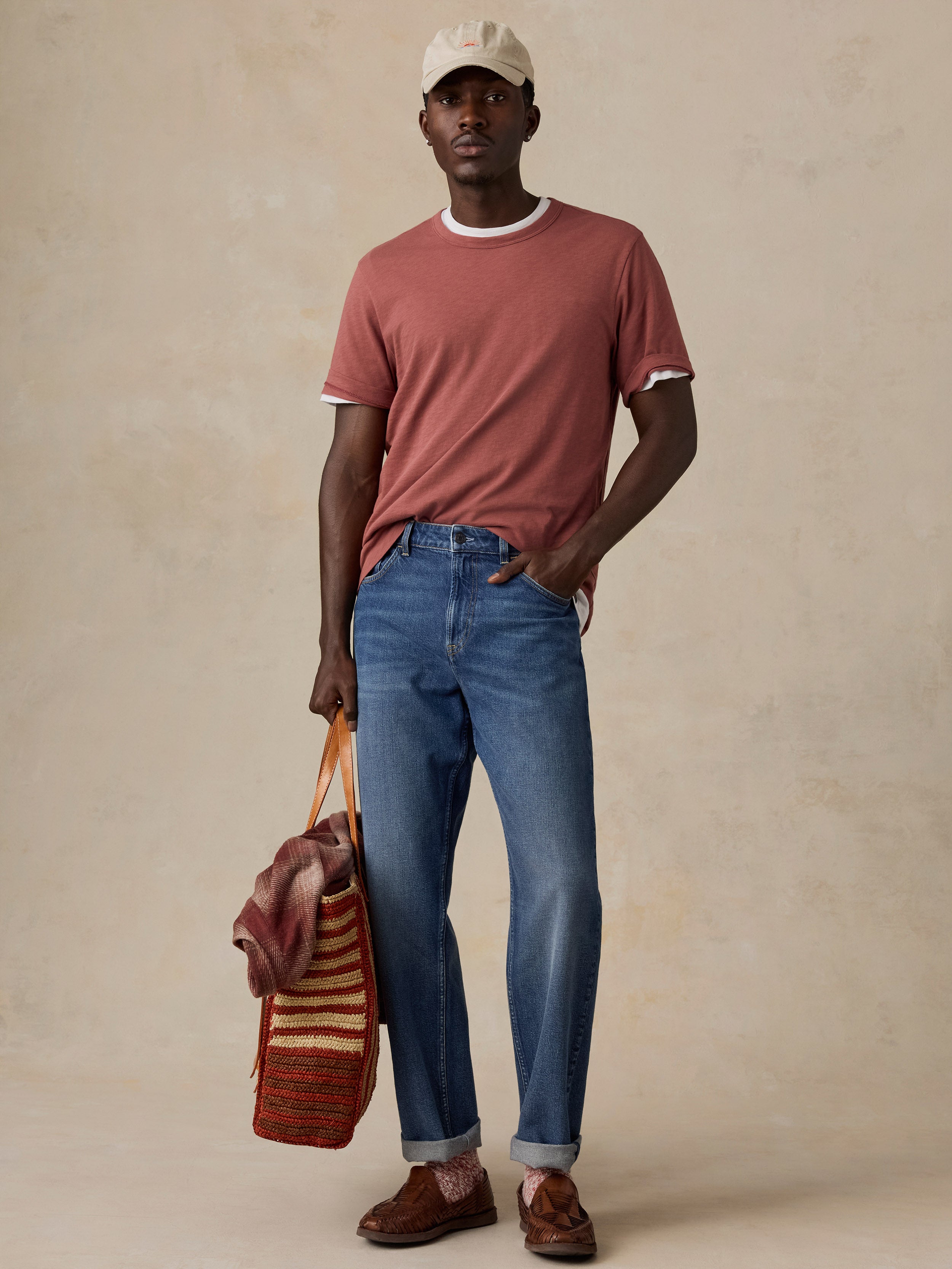 A man modeling a dusty rose ringer t-shirt with white trim, blue jeans, brown shoes, and a beige cap, holding a striped knit bag against a textured beige backdrop.