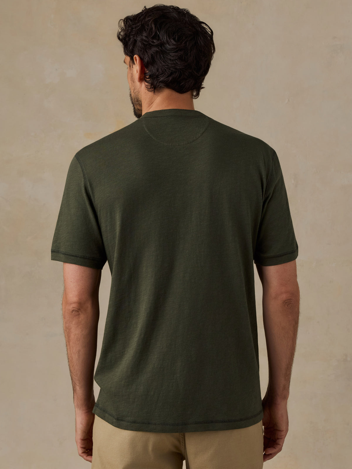 A man viewed from behind wearing an olive green short-sleeve crewneck t-shirt and khaki shorts, standing against a textured beige wall., image 3 of 6