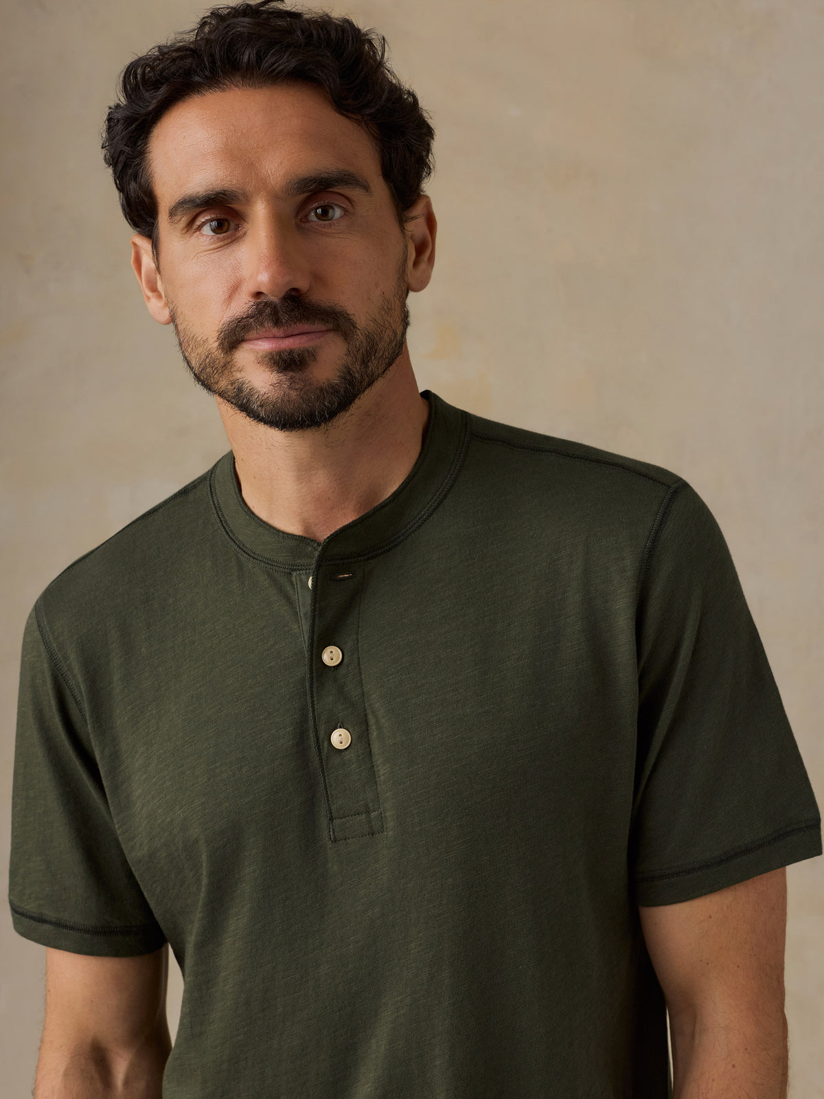 A man wearing a dark olive green short-sleeve henley shirt with a three-button placket, photographed against a neutral textured beige background., image 2 of 6
