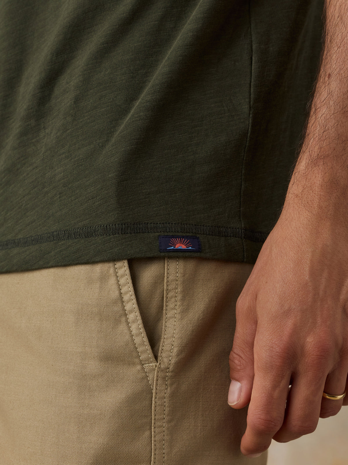 Close-up of a man wearing an olive green short-sleeve t-shirt and khaki shorts, showing a small Faherty brand logo patch on the hem., image 4 of 6