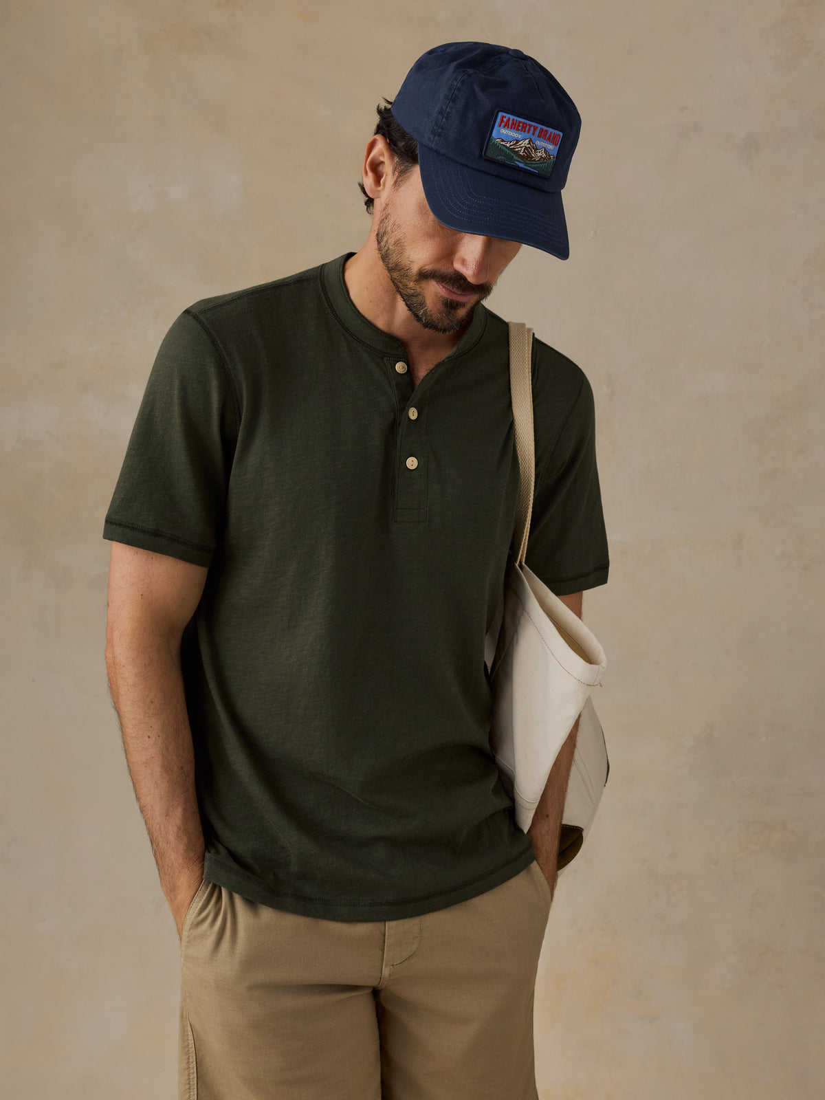 A man wearing a dark olive green short-sleeve henley shirt, khaki shorts, a navy blue cap, and carrying a cream canvas shoulder bag, set against a textured beige studio backdrop., image 5 of 6