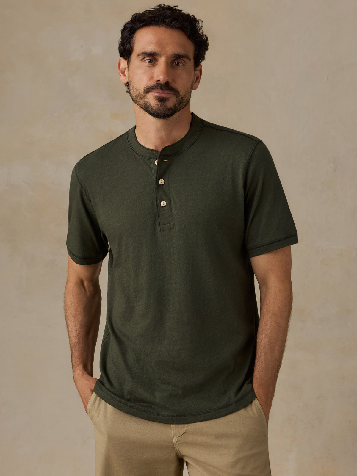 A man wearing a dark olive green short-sleeve henley shirt with three buttons, paired with khaki shorts, standing against a textured beige studio background., image 1 of 6