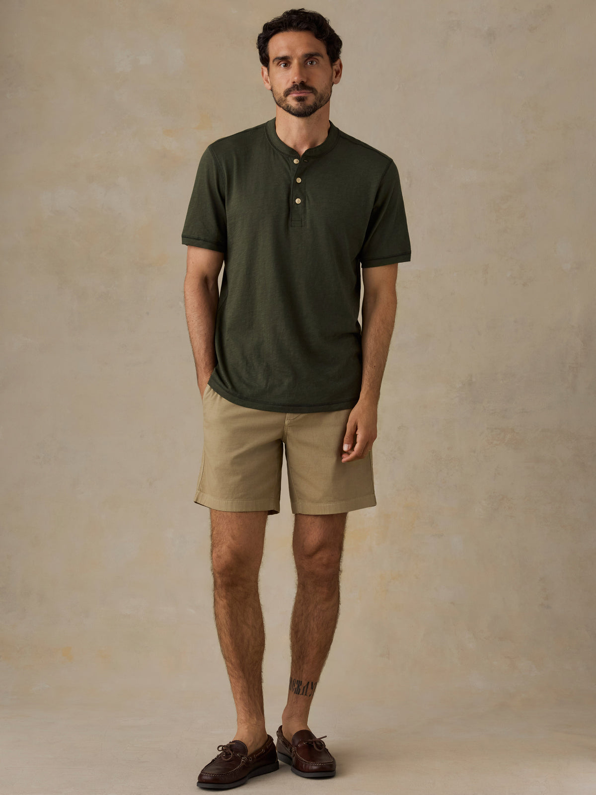 A man wearing an olive green short-sleeve henley shirt, beige shorts, and dark brown loafers, standing against a textured beige studio backdrop., image 6 of 6