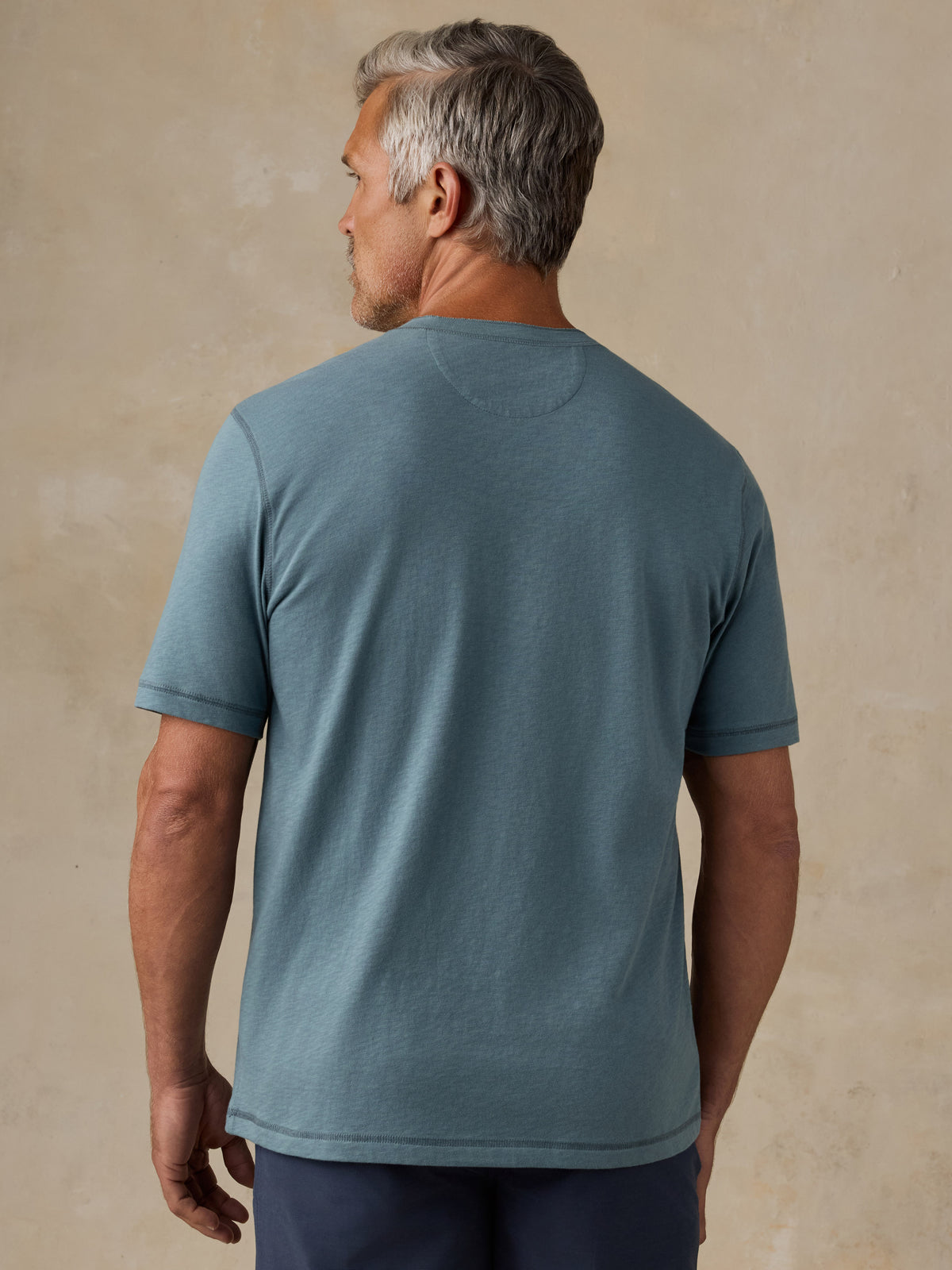 A man viewed from the back wearing a muted slate blue crewneck t-shirt and dark navy pants, standing against a textured beige studio backdrop., image 3 of 6
