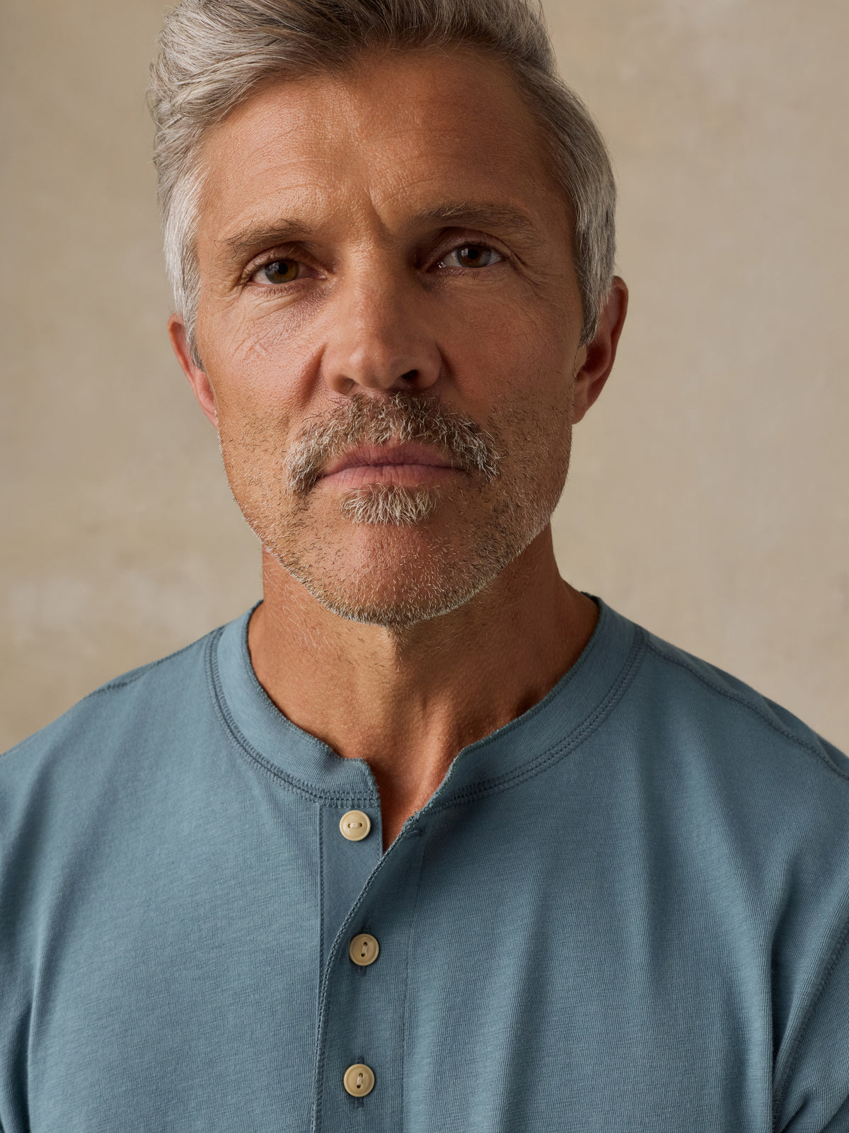 A close-up portrait of a man with gray hair and a mustache wearing a dusty blue henley shirt with light-colored buttons, set against a neutral beige background., image 2 of 6