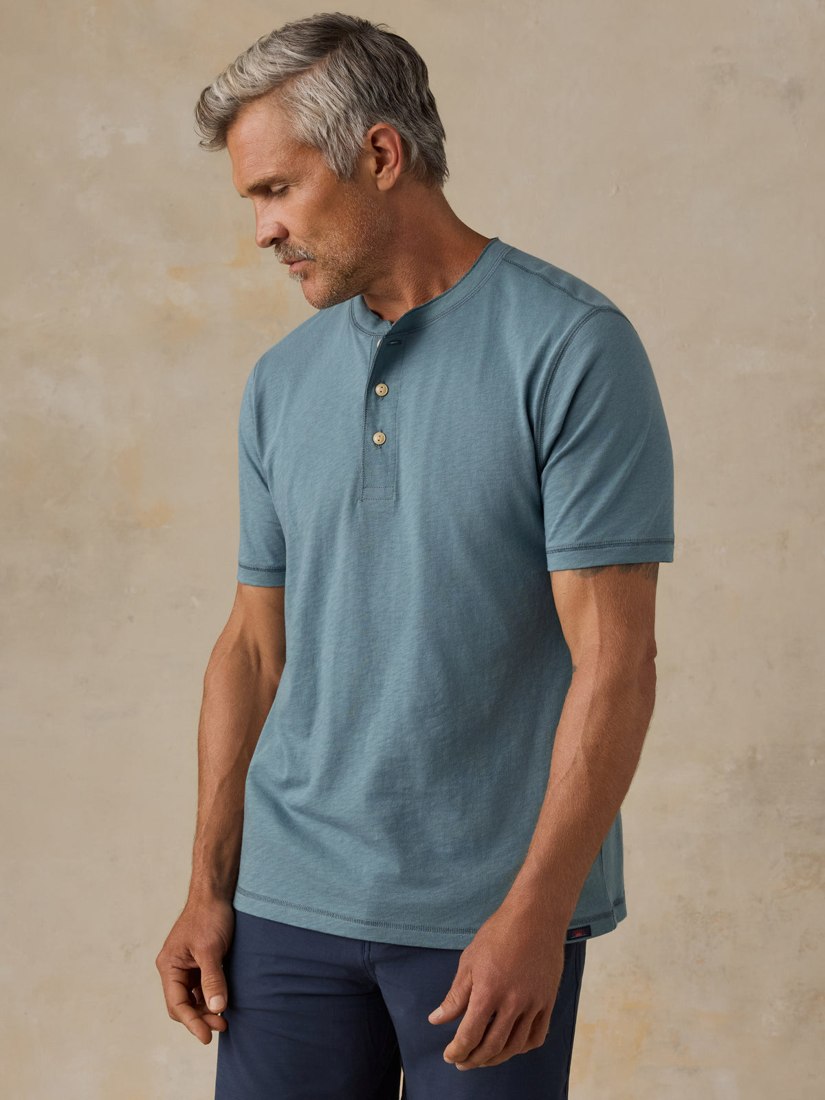A man wearing a muted blue short-sleeve henley shirt with a three-button placket, standing against a textured beige studio backdrop., image 1 of 6