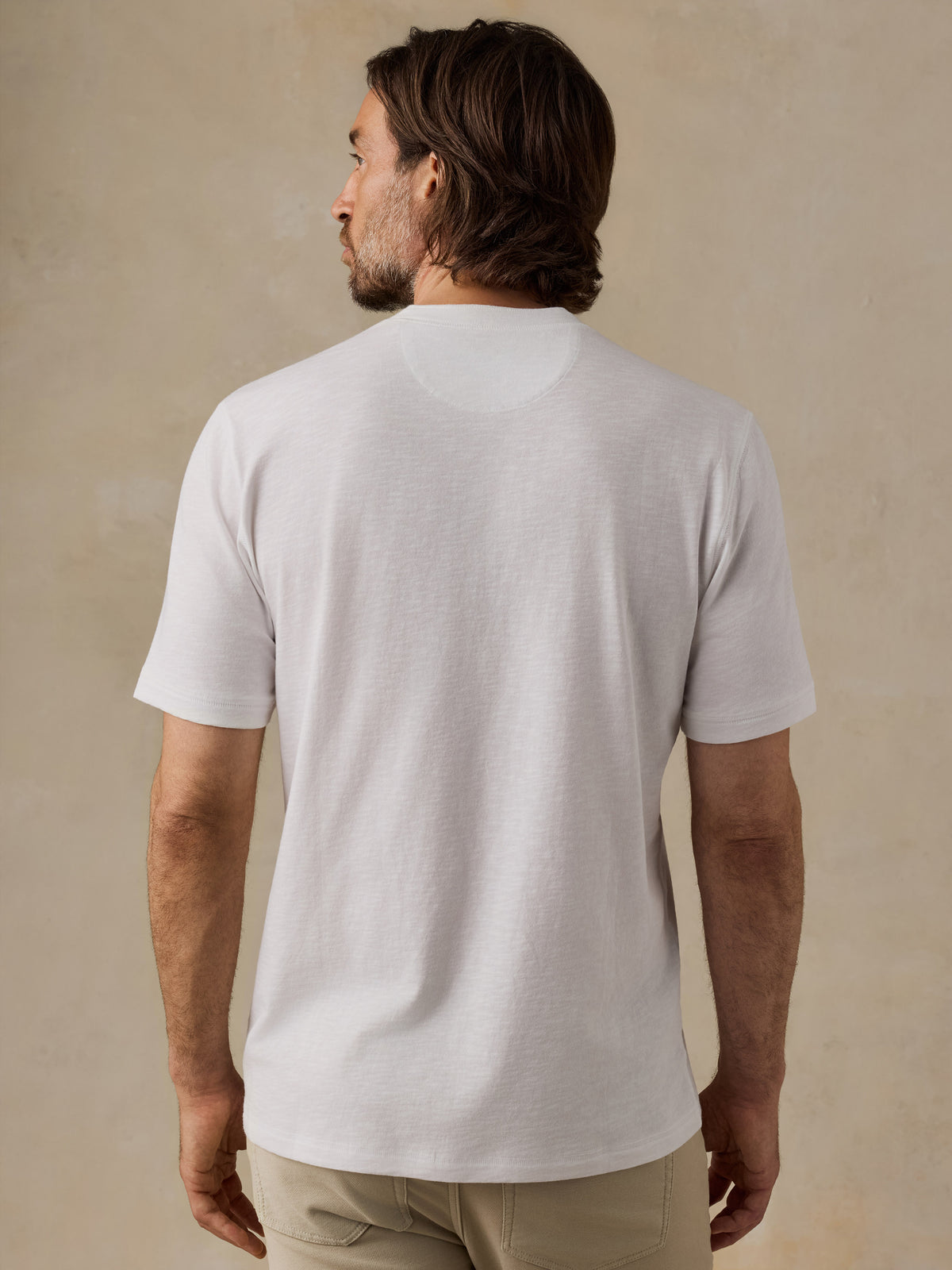 A man viewed from the back wearing a plain white short-sleeve crewneck t-shirt and khaki pants, standing against a textured beige studio wall., image 3 of 6