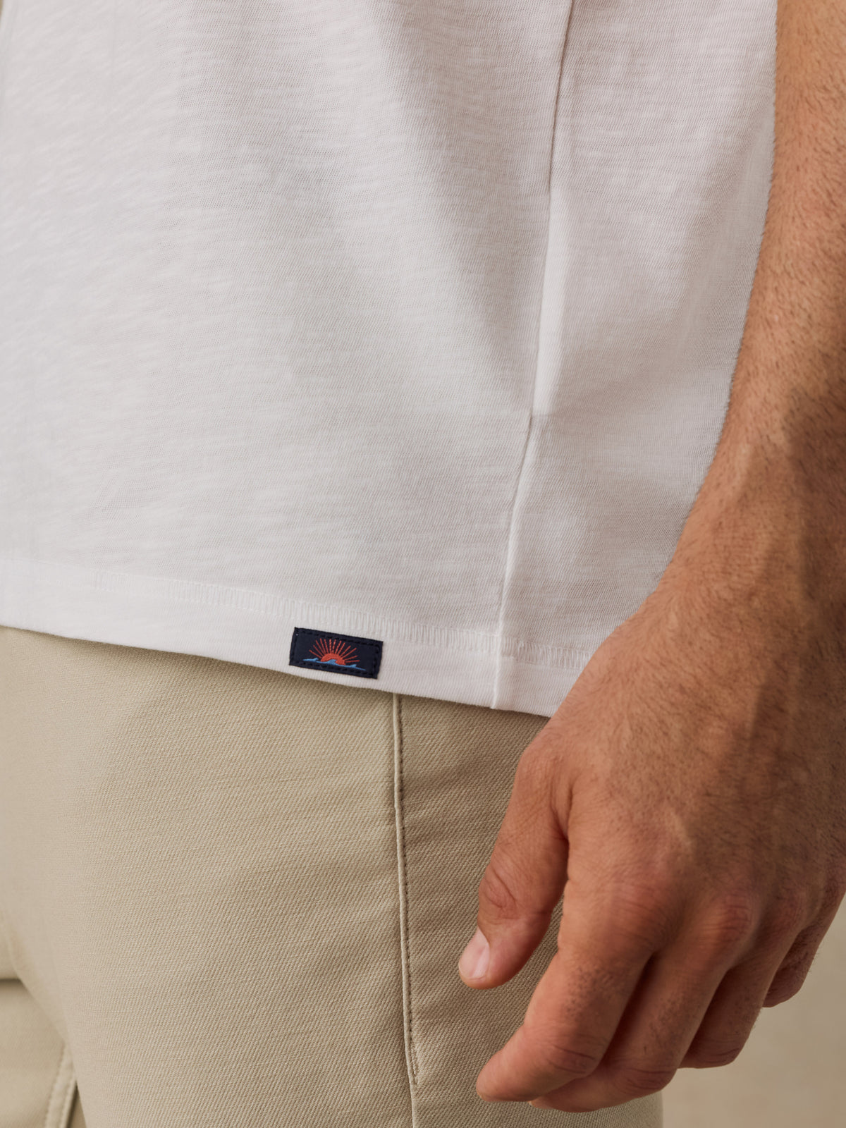 Close-up of a man wearing a white short-sleeve t-shirt and beige trousers, with a small navy Faherty brand patch on the hem of the shirt., image 4 of 6