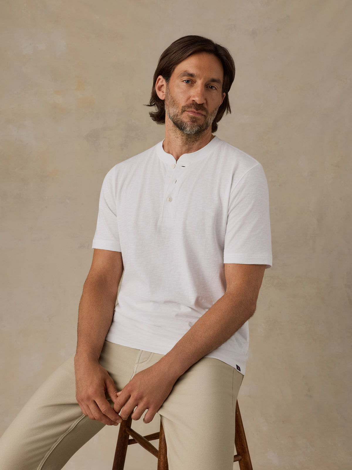 A man wearing a white short-sleeve henley shirt and beige trousers, sitting on a wooden stool against a textured beige studio backdrop., image 6 of 6
