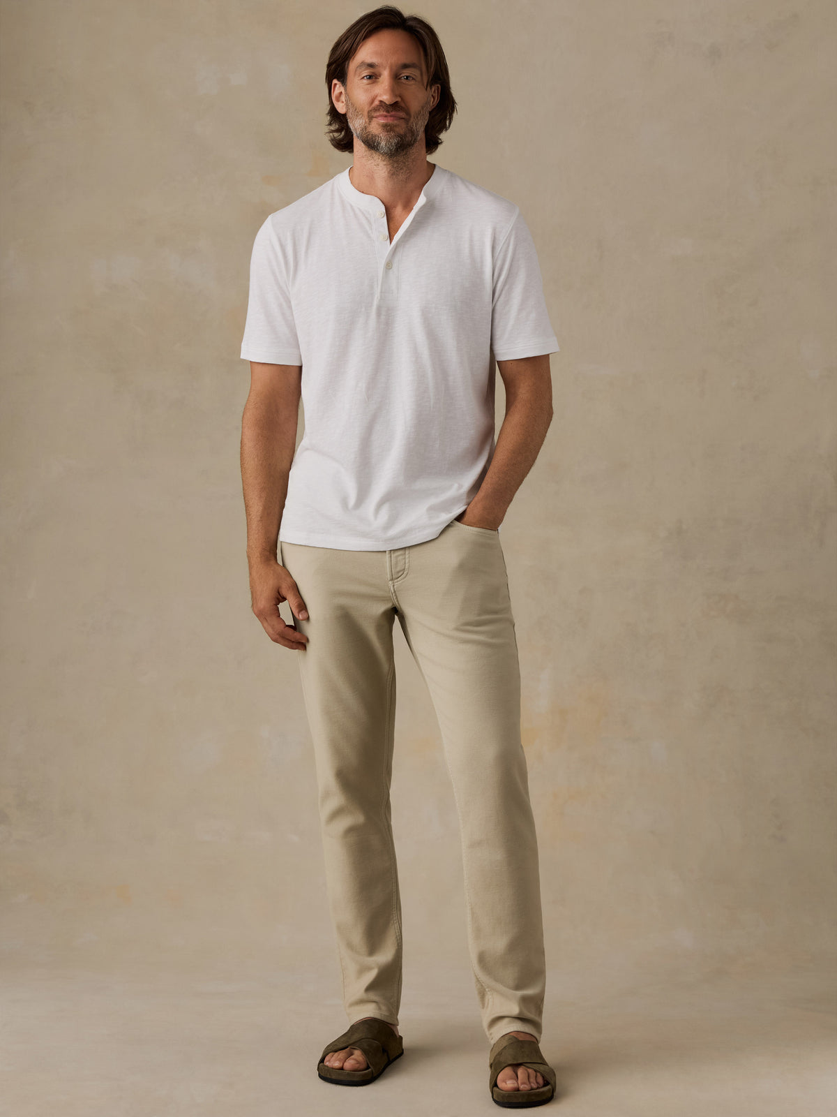 A man wearing a white short-sleeve henley shirt, beige trousers, and brown sandals, standing against a textured beige studio backdrop., image 5 of 6