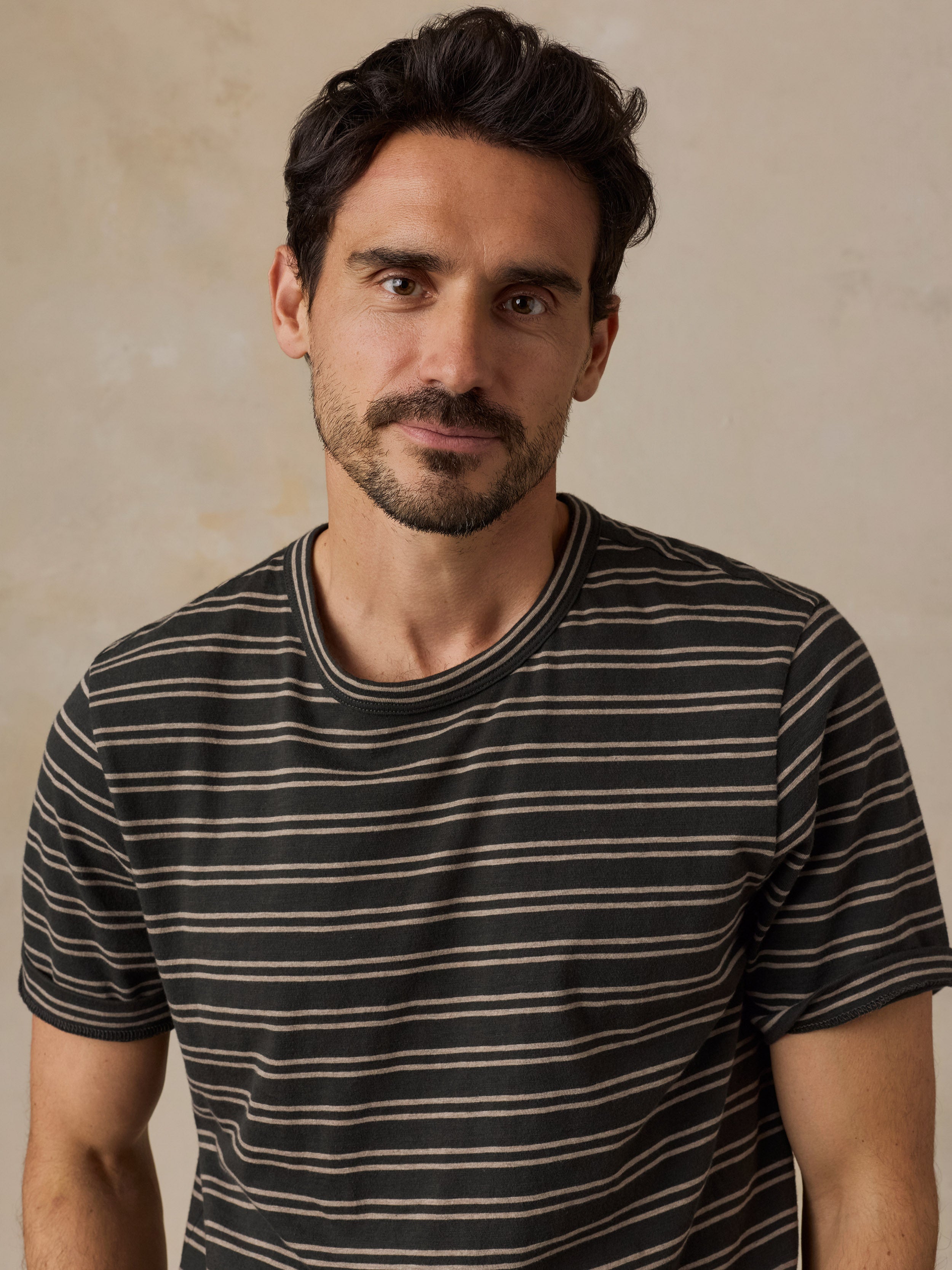 A man wearing a short-sleeve crewneck t-shirt with thin horizontal stripes in black and beige, set against a neutral textured beige background.