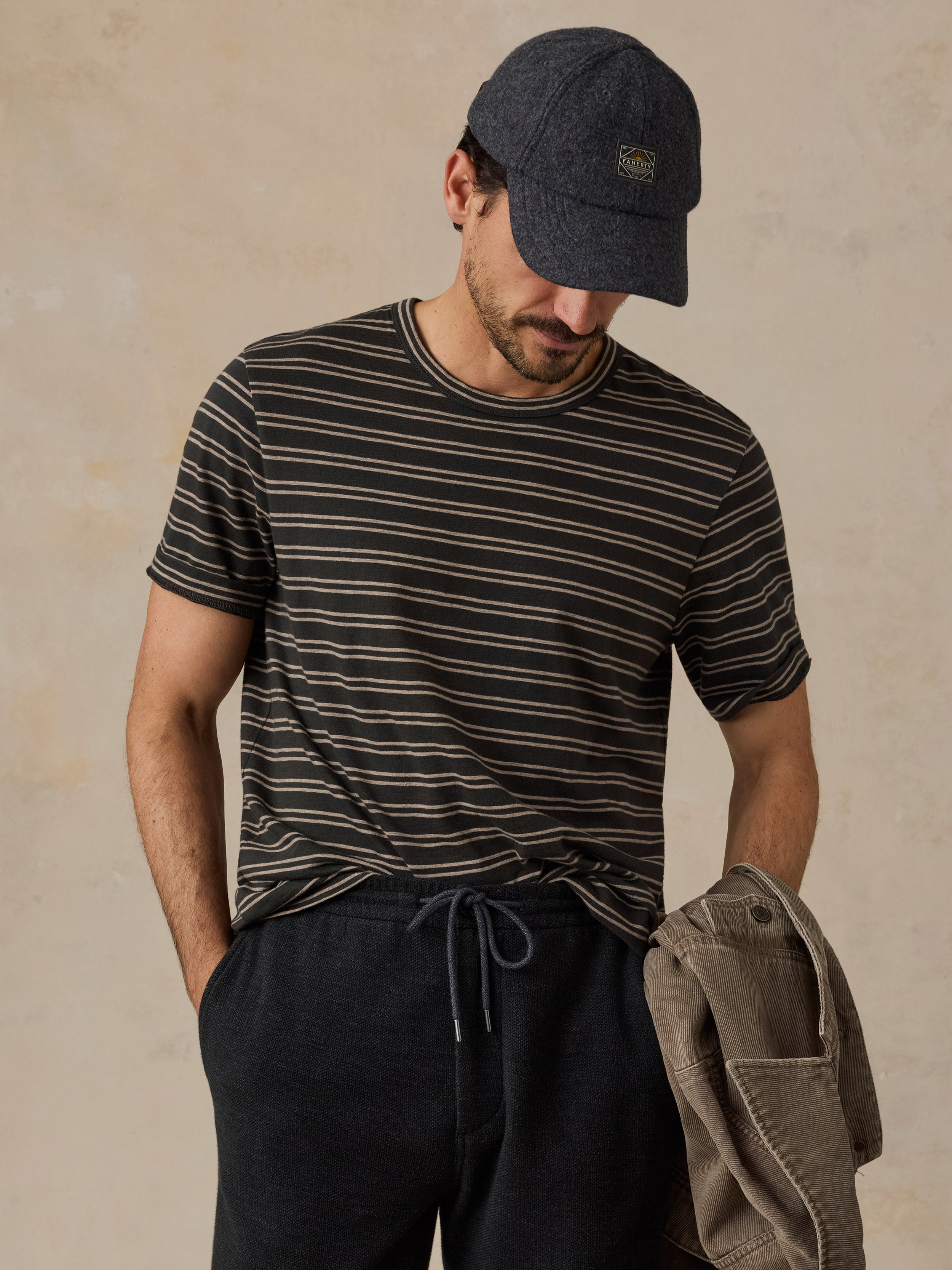 A man wearing a black and beige horizontally striped t-shirt, a dark navy baseball cap, and dark charcoal drawstring sweatpants, holding a beige jacket against a textured beige background.