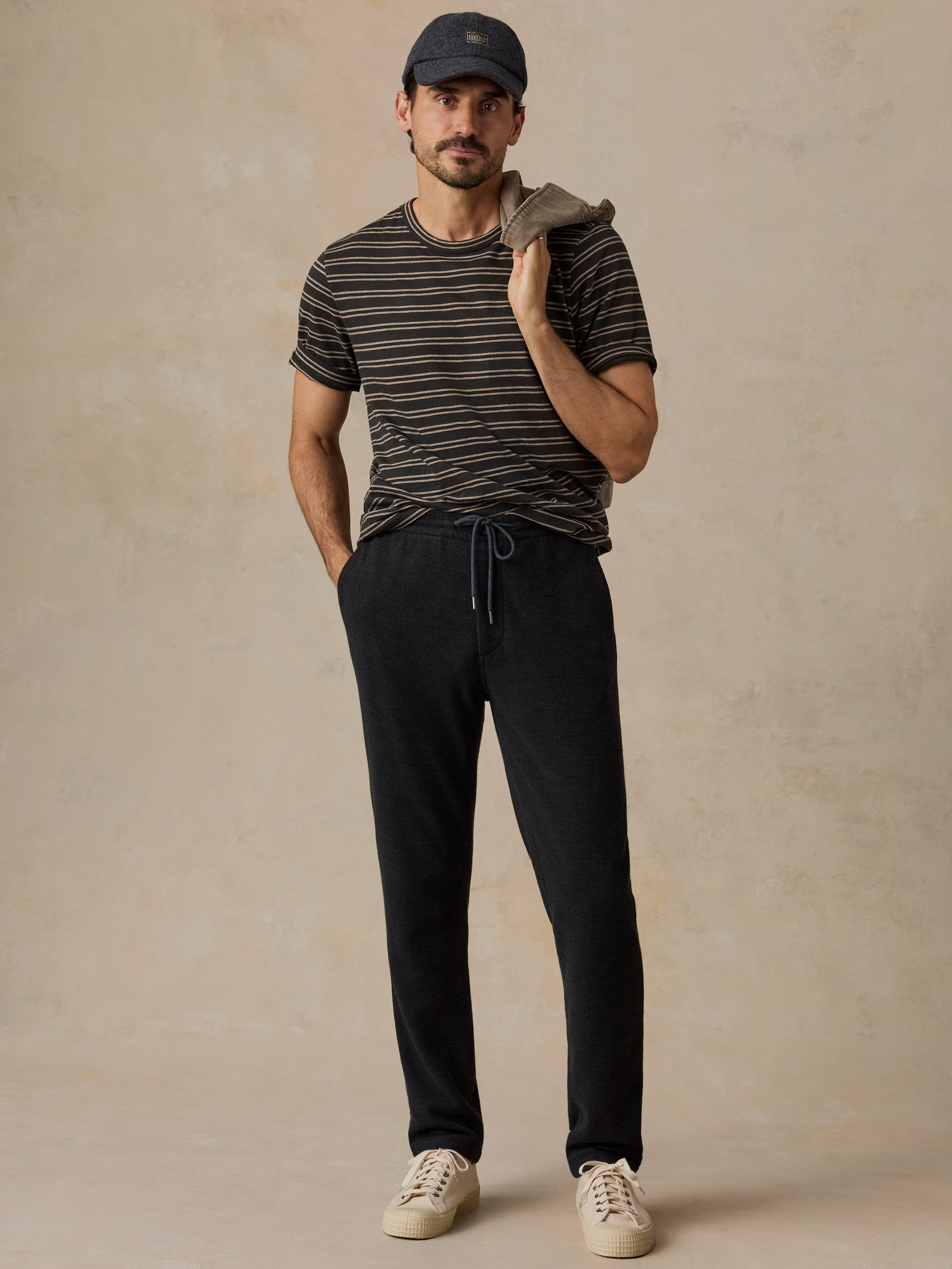A man modeling a black and beige striped t-shirt, black drawstring sweatpants, a dark cap, and off-white sneakers, standing against a textured beige studio backdrop.