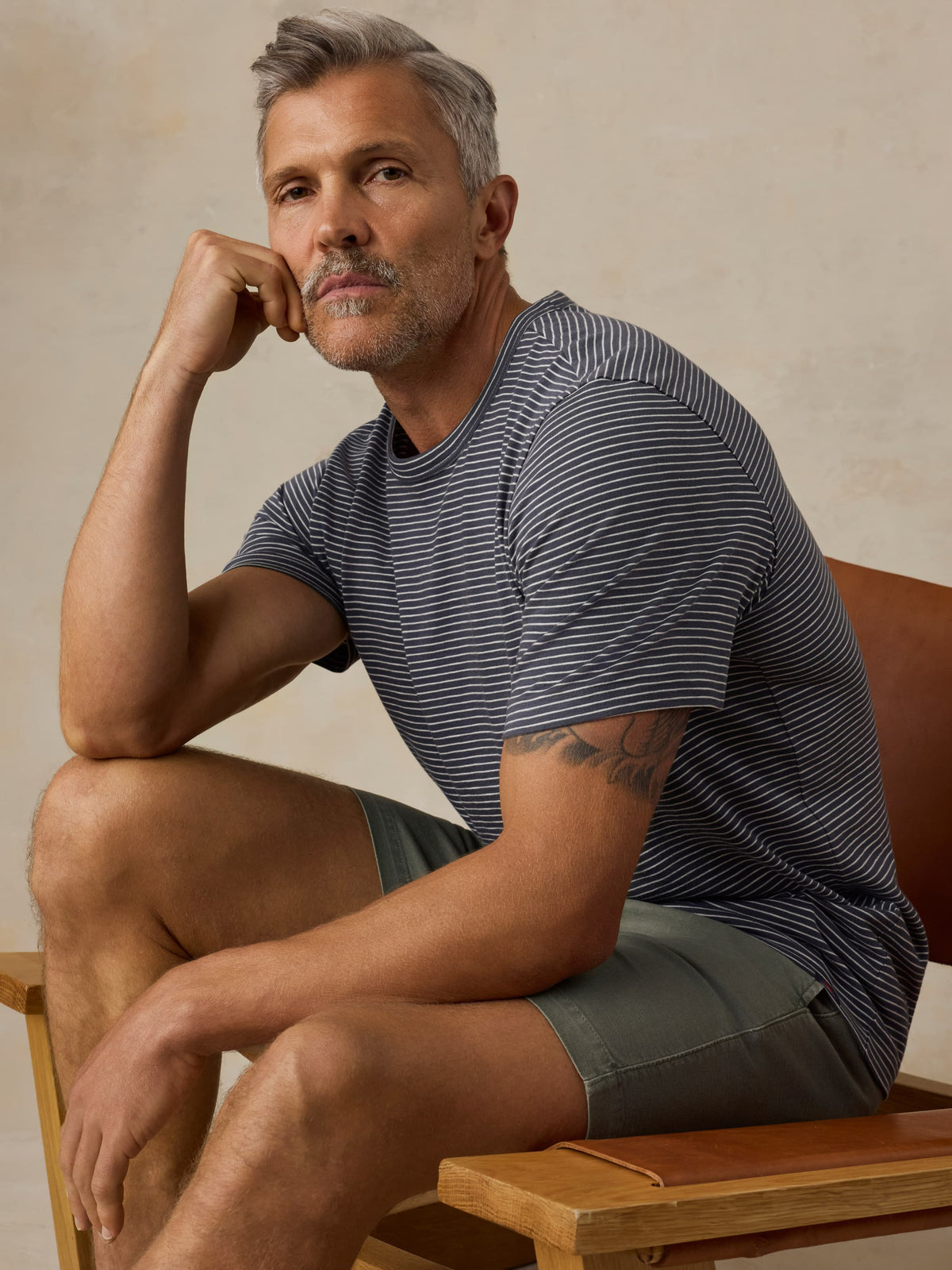 Man sitting on a wooden chair wearing blue striped shirt and green shorts against a beige background, image 2 of 5