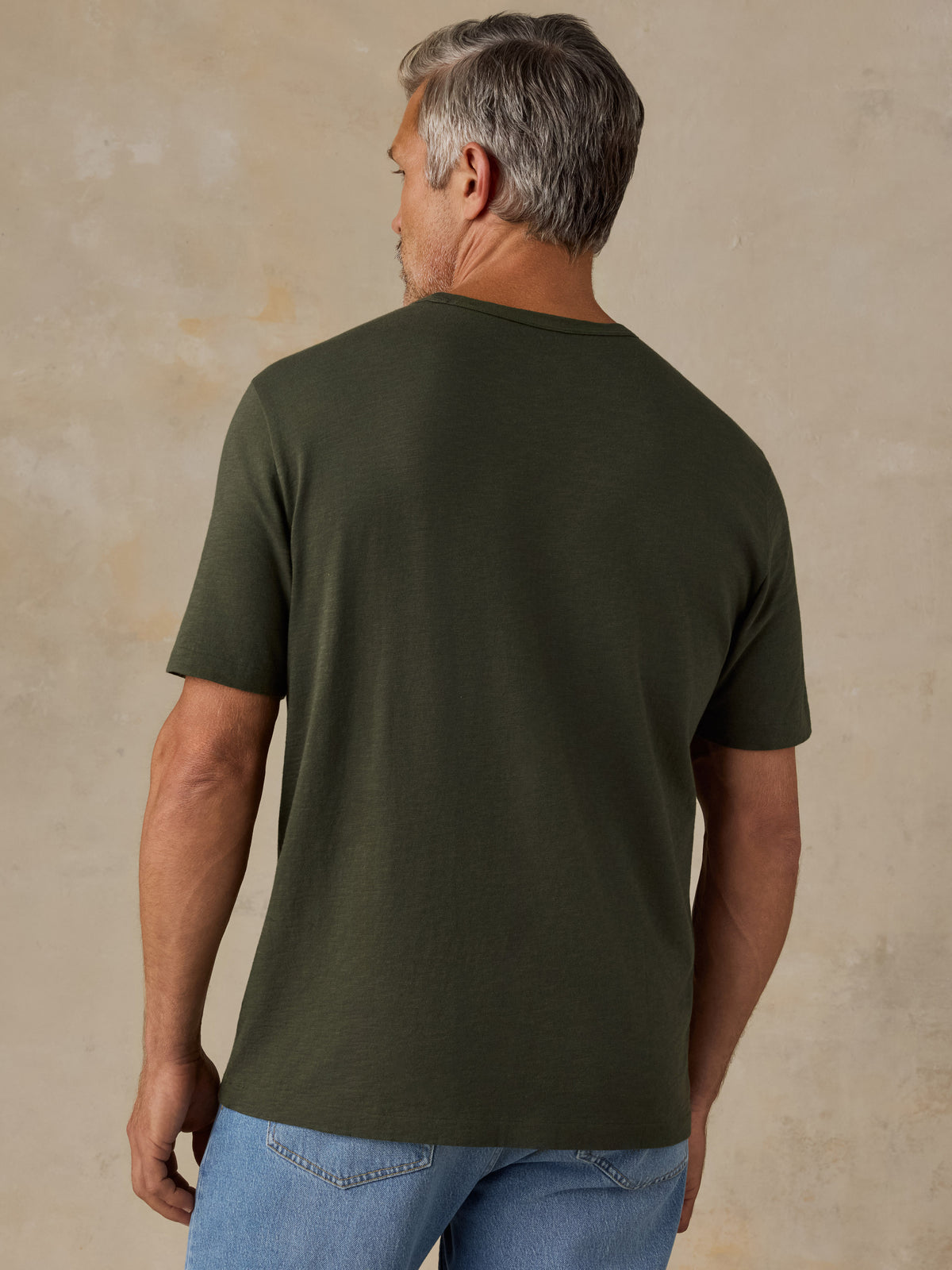 A man viewed from the back wearing a solid olive green short-sleeve t-shirt and blue jeans, standing against a textured beige studio wall., image 3 of 8