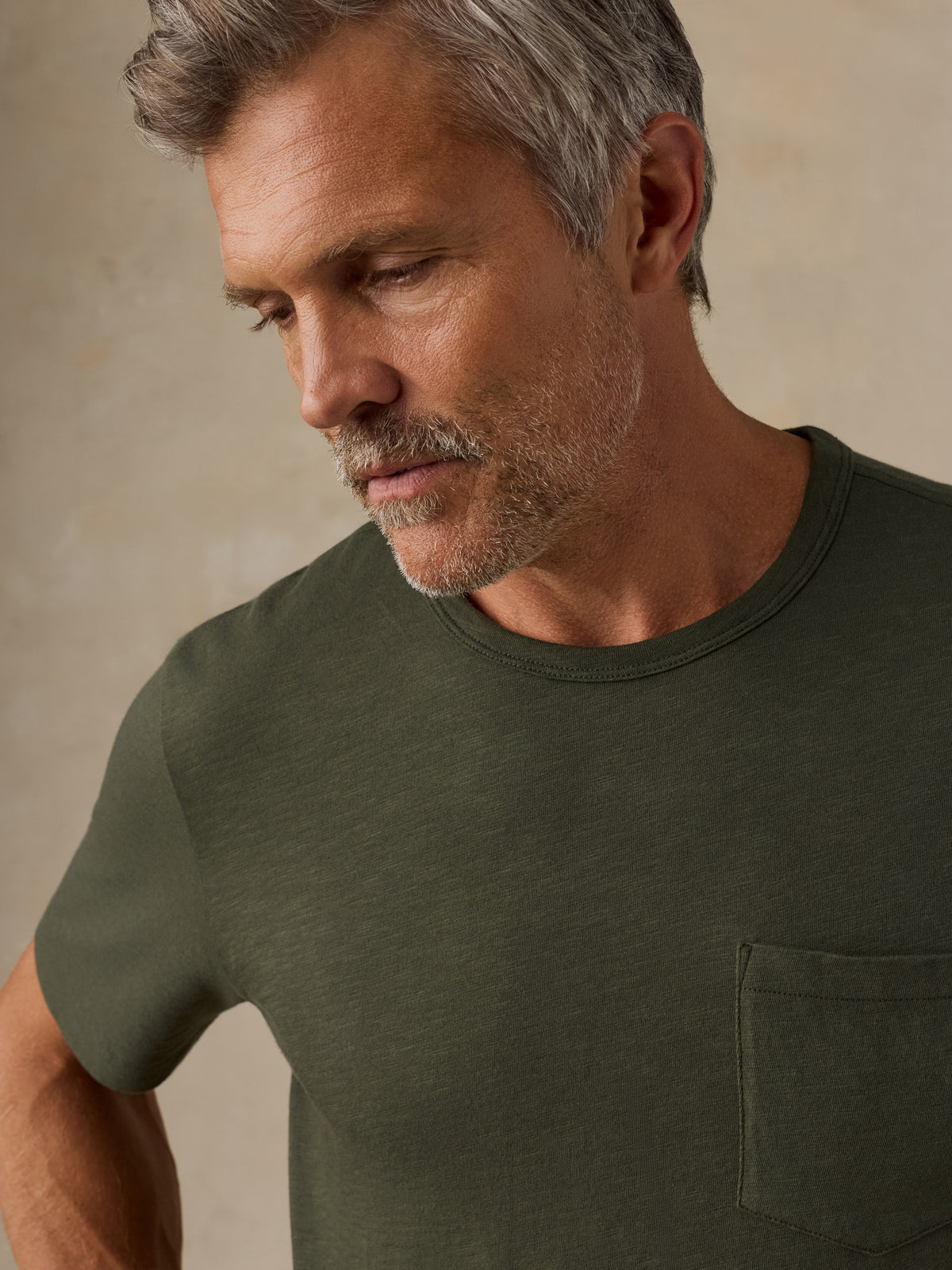 A close-up of a man wearing a dark olive green crew-neck t-shirt with a chest pocket, set against a neutral beige background., image 2 of 8