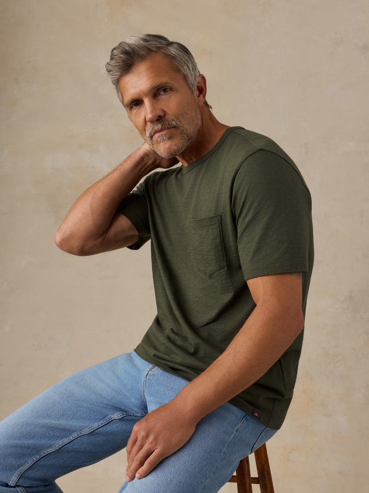 A man with gray hair and a beard sits on a wooden stool wearing an olive green short-sleeve pocket t-shirt and light blue jeans against a textured beige background., image 5 of 8