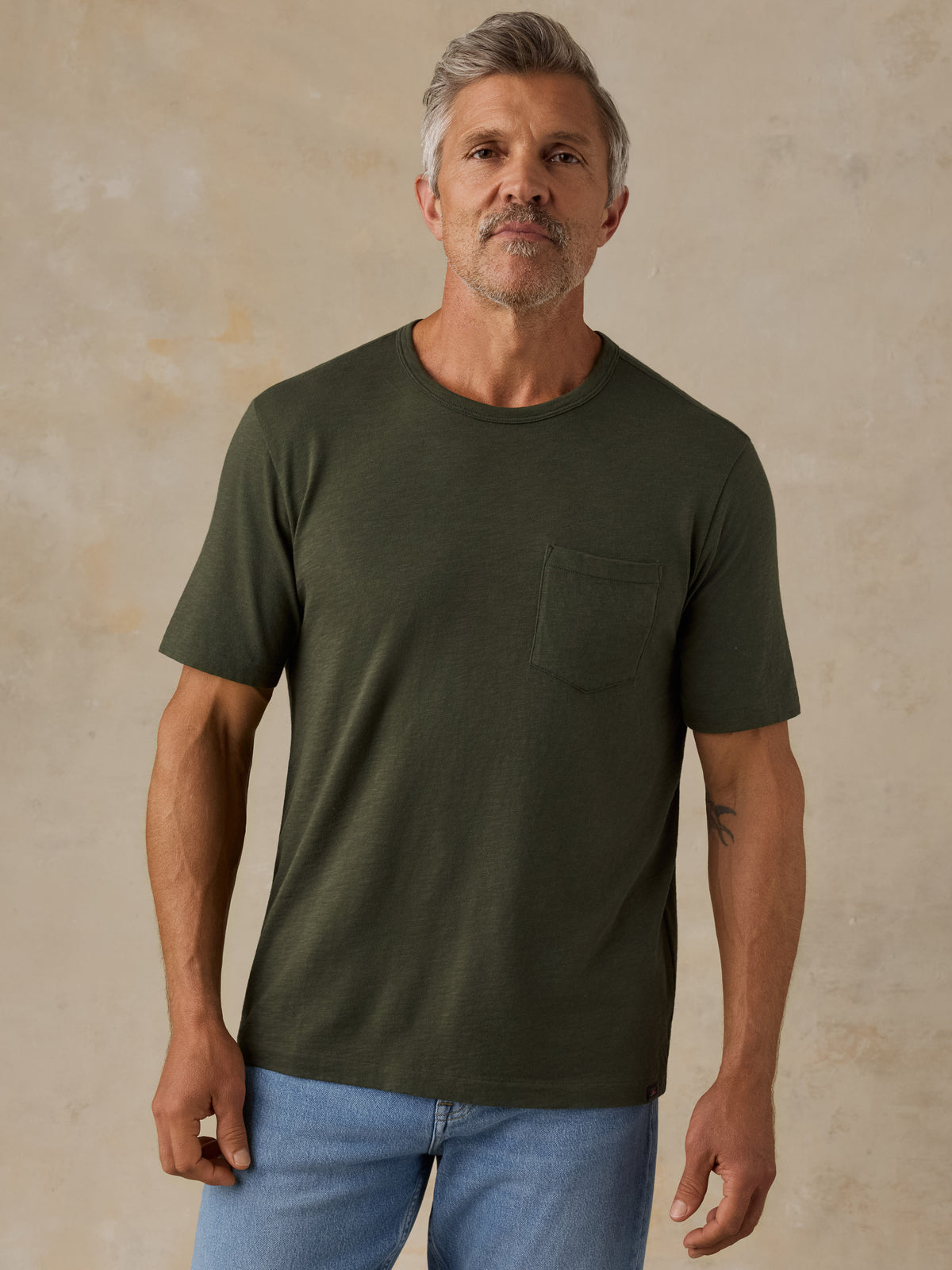 A man wearing a dark olive green short-sleeve crewneck t-shirt with a chest pocket, paired with light blue jeans, standing against a textured beige wall., image 1 of 8