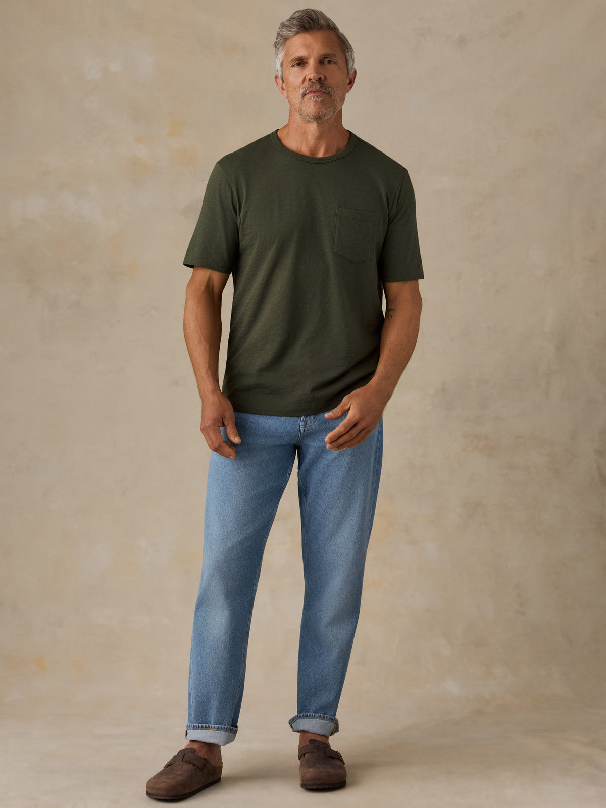 A man wearing an olive green short-sleeve crewneck pocket t-shirt, light blue jeans, and brown suede mules, standing against a textured beige studio backdrop., image 6 of 8