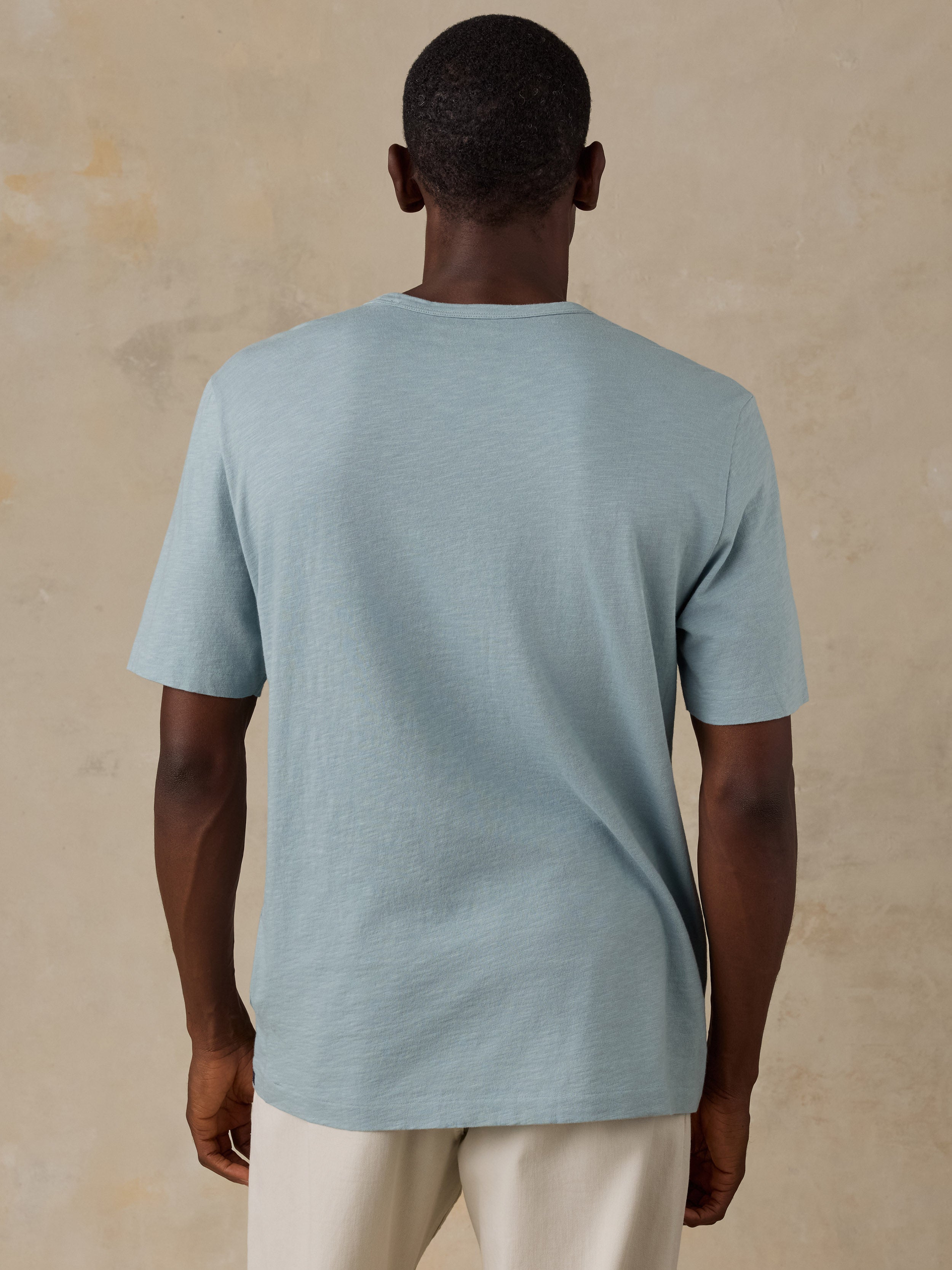 A man viewed from behind wearing a light blue short-sleeve crewneck t-shirt and cream-colored pants, standing against a textured beige background.