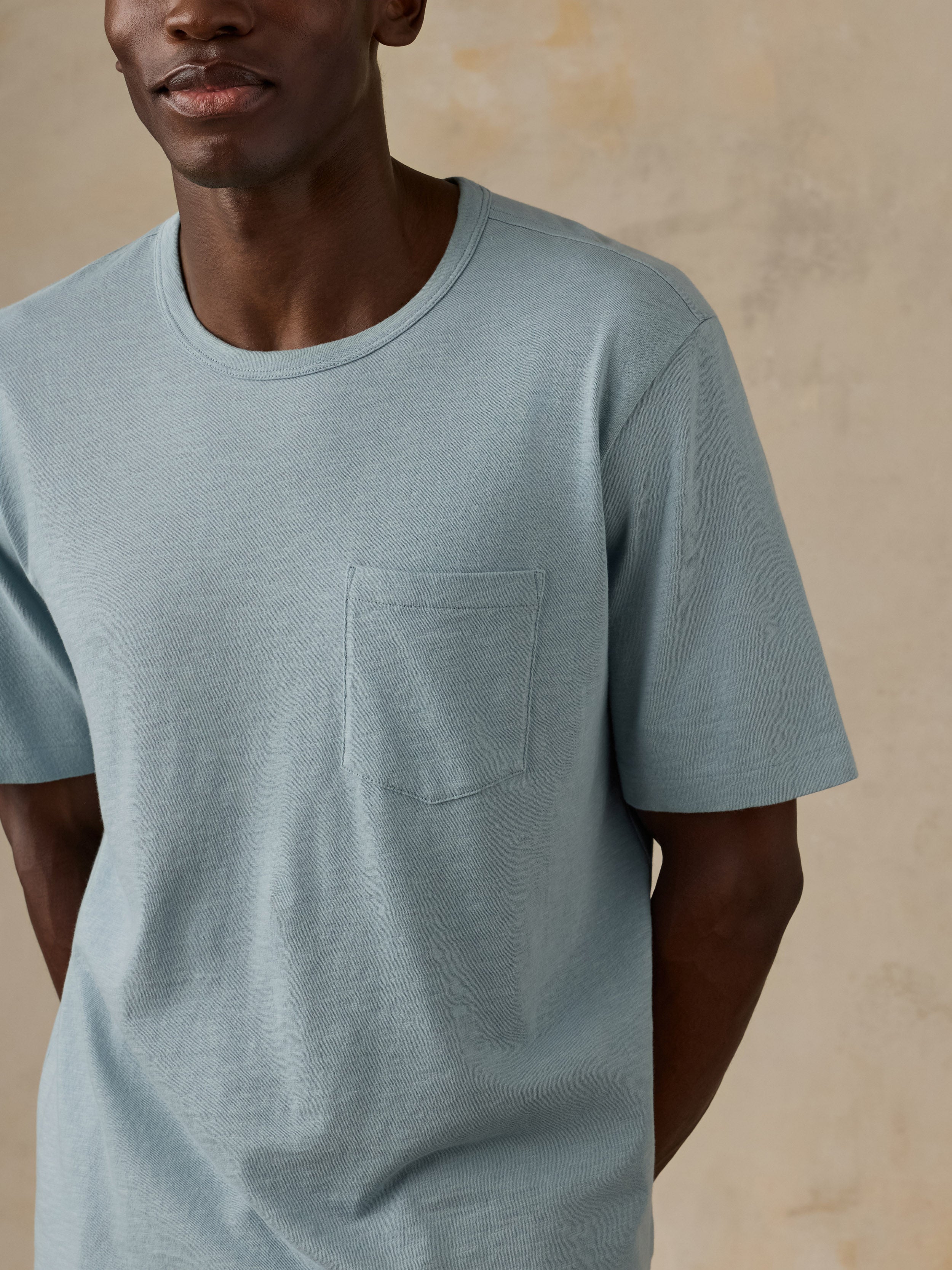 A close-up of a man wearing a light blue crewneck t-shirt with a chest pocket, set against a textured beige background.