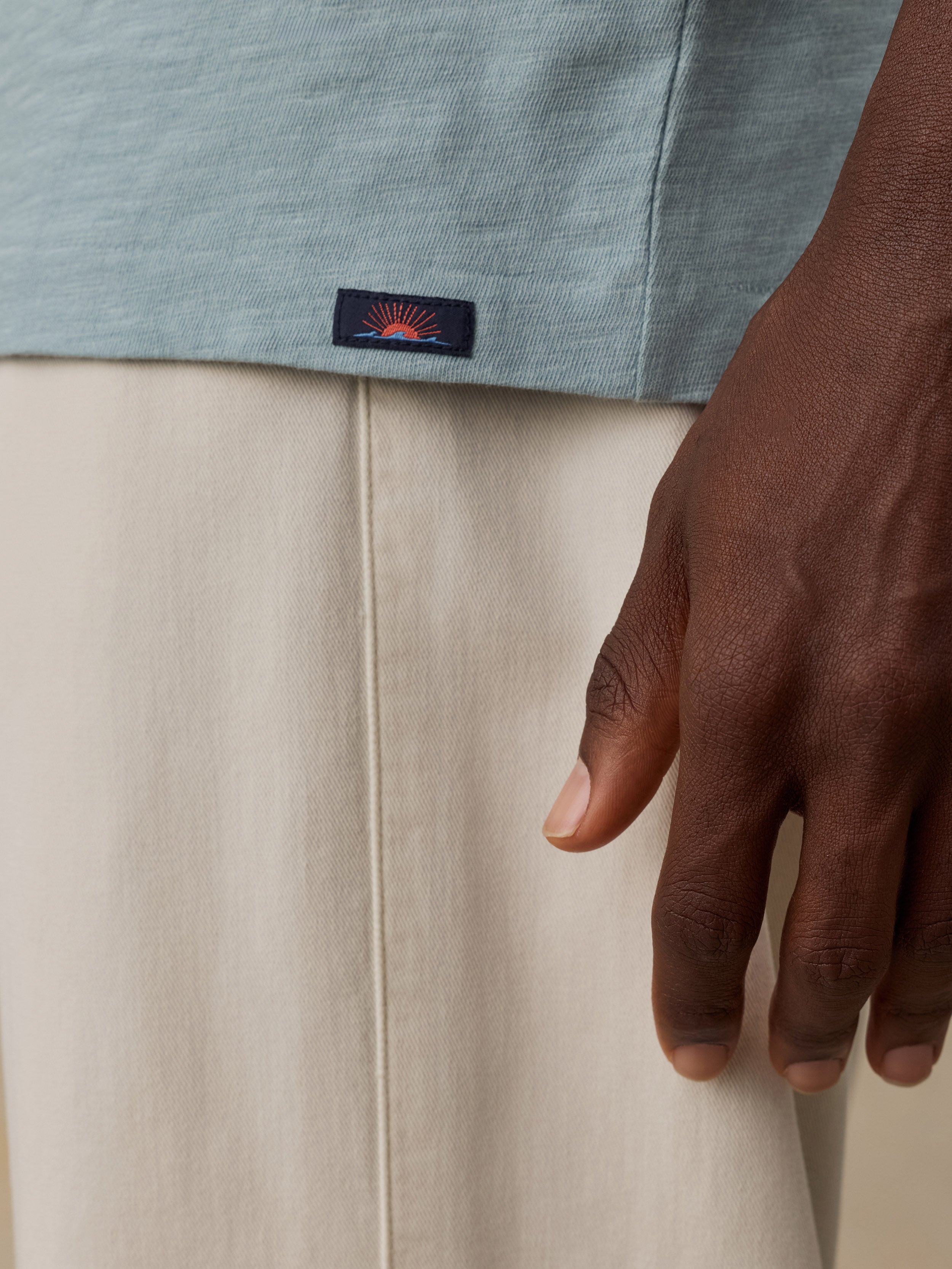 Close-up of a man wearing a light blue-gray heathered top and cream-colored pants, with a small rectangular Faherty brand patch visible near the hem of the top.