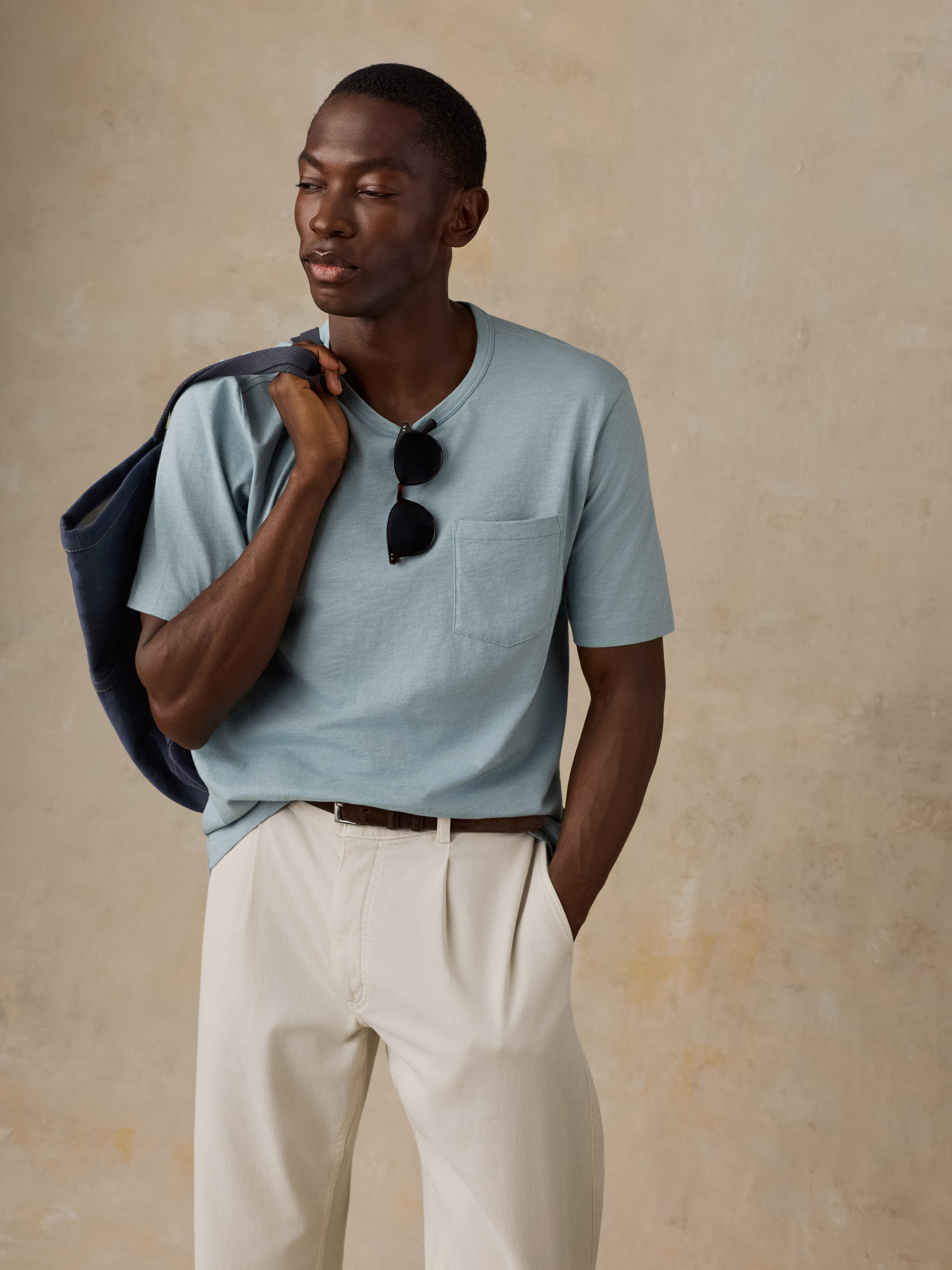 A man wearing a light blue short-sleeve pocket t-shirt, cream-colored pleated trousers, and carrying a dark blue tote bag over his shoulder, set against a textured beige background.