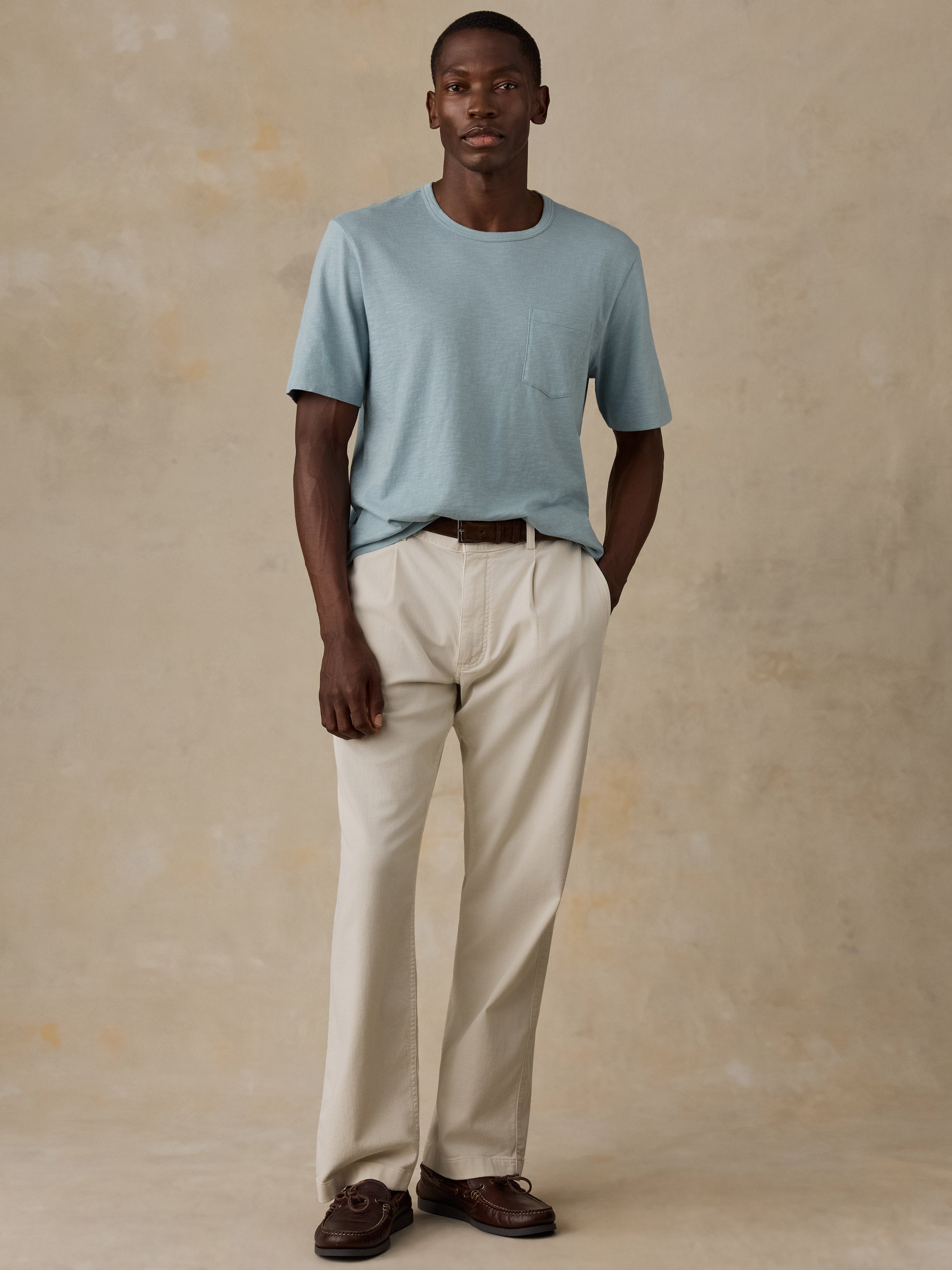 A man modeling a light blue crew-neck t-shirt and cream-colored trousers, standing against a textured beige studio backdrop.