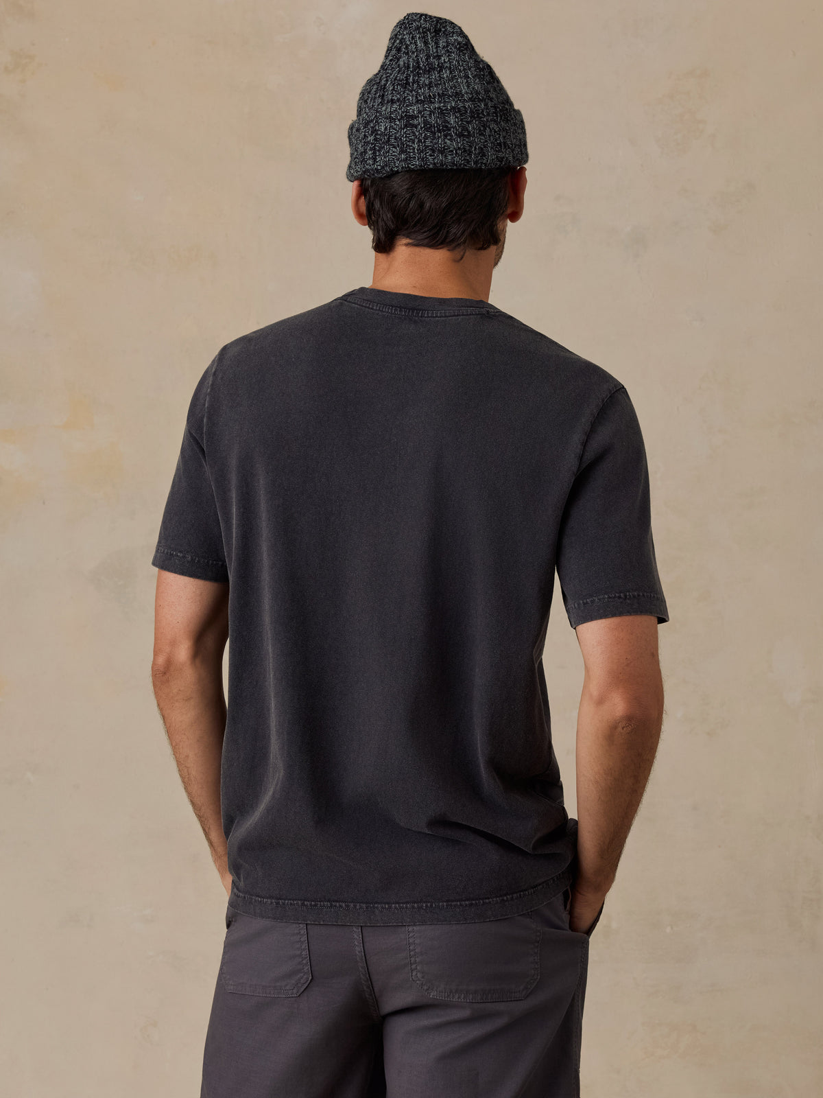 A man viewed from behind wearing a dark charcoal gray short-sleeve t-shirt, dark gray pants, and a dark gray knit beanie, standing against a textured beige wall., image 3 of 6