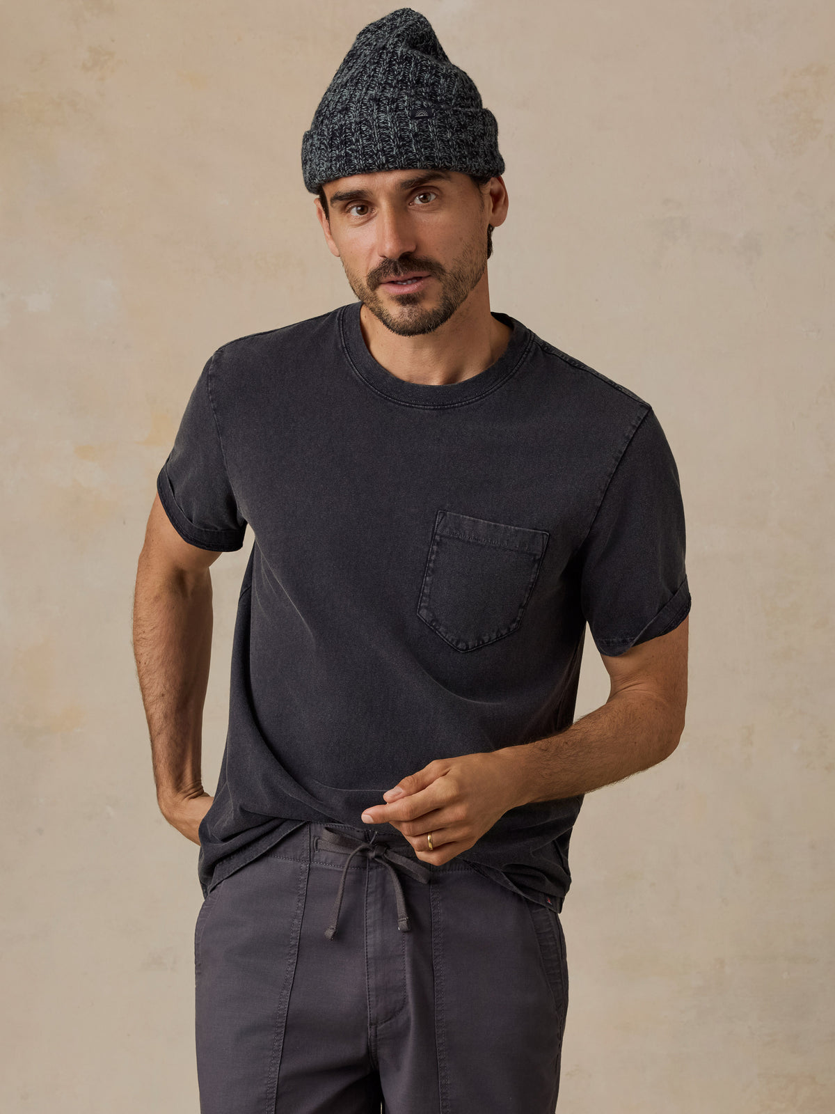 A man wearing a dark charcoal gray crewneck pocket t-shirt, matching dark gray drawstring pants, and a dark gray knit beanie, standing against a textured beige studio background., image 1 of 6