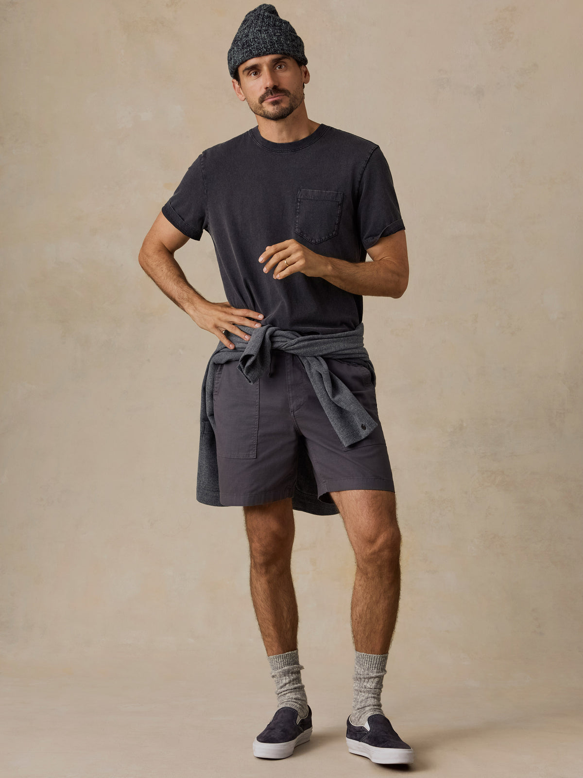 A man modeling a dark charcoal gray t-shirt with a chest pocket, matching dark gray shorts, a gray beanie, and gray slip-on shoes against a textured beige studio backdrop., image 5 of 6