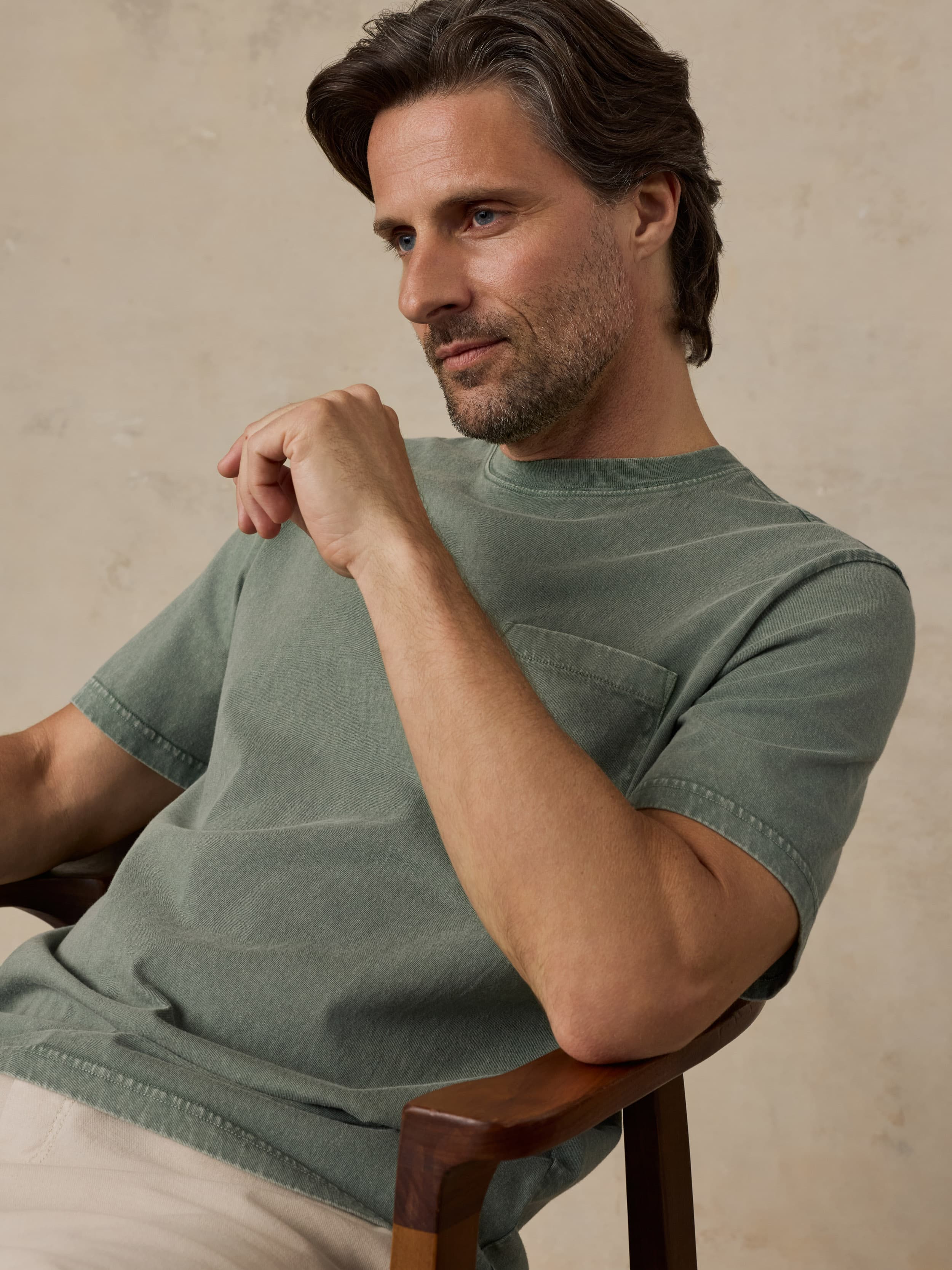 Man wearing a green t-shirt sitting on a wooden chair against a beige background