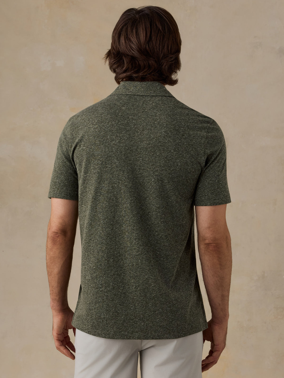 A man viewed from the back wearing a short-sleeve knit polo shirt in heathered olive green, paired with light-colored pants, against a textured beige studio backdrop., image 3 of 6