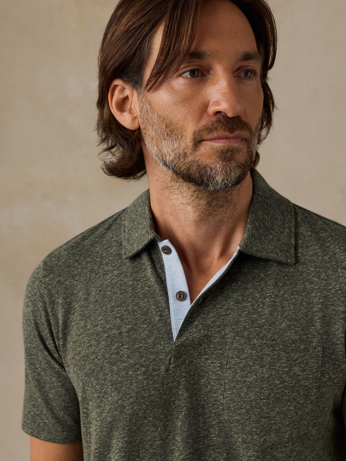 A man wearing a short-sleeve knit polo shirt in heathered olive green, featuring a light blue inner placket and dark buttons, set against a neutral beige background., image 2 of 6