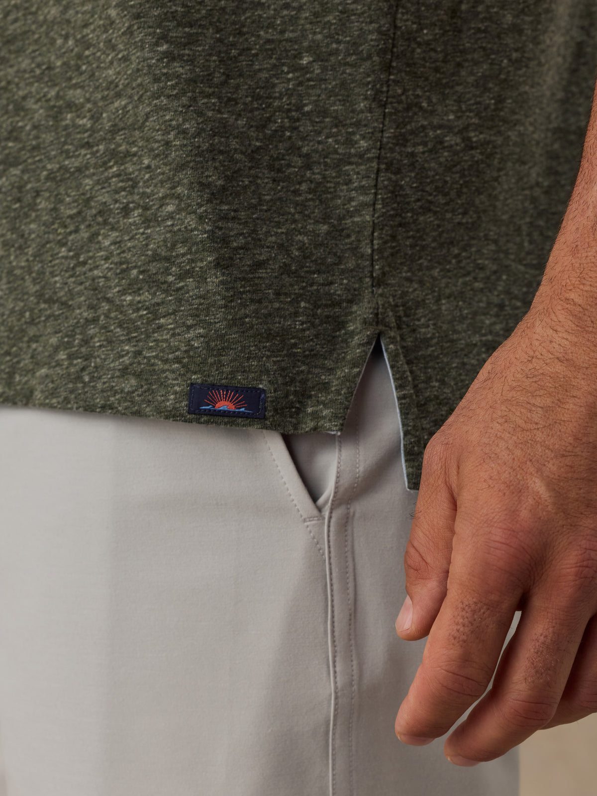 Close-up of a man wearing a dark heathered green knit top and light gray pants, with a small Faherty brand patch near the hem., image 5 of 6